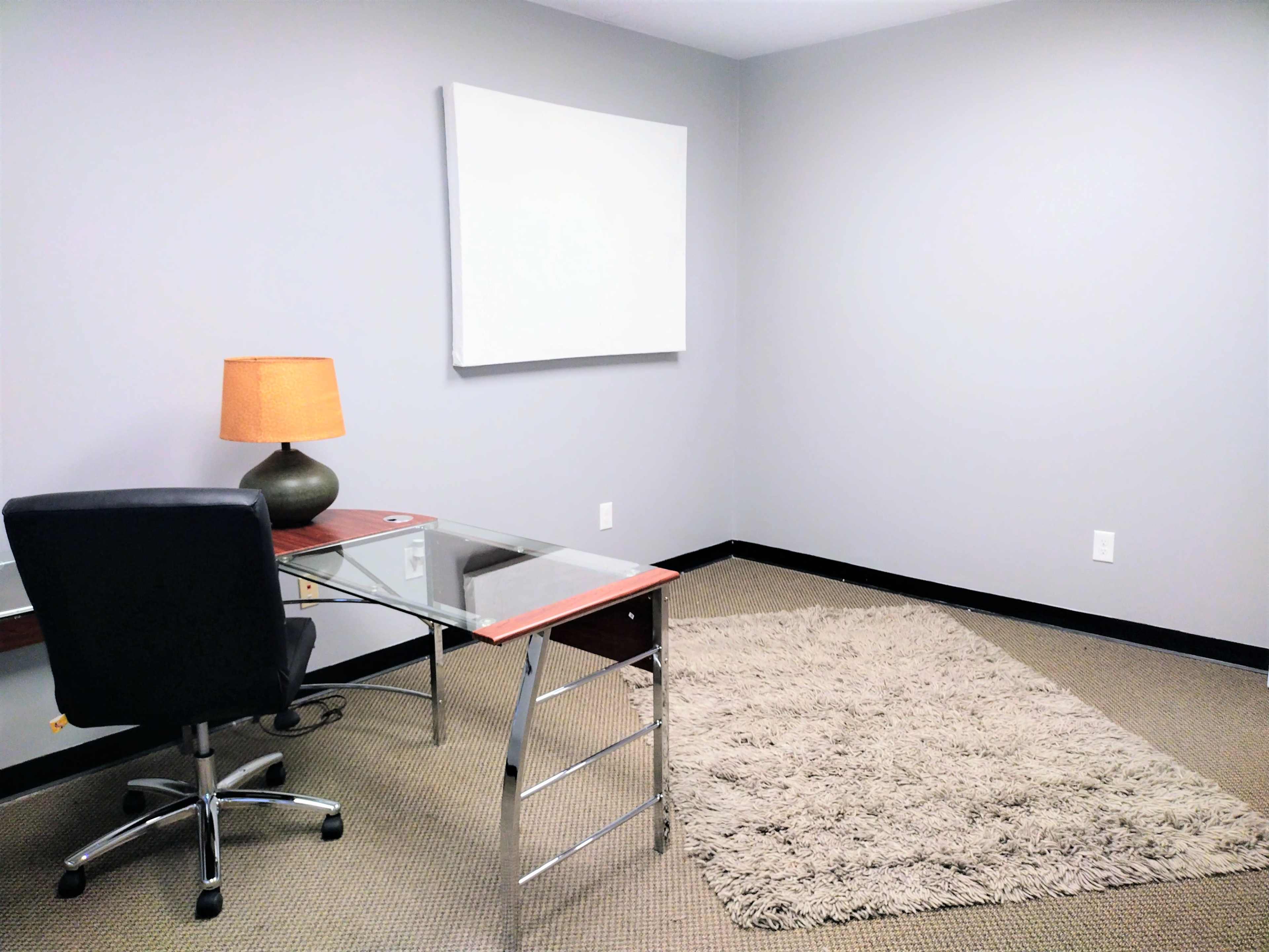 A minimalistic office space with a glass-top desk, an ergonomic chair, a lamp, and a large area rug on the floor.