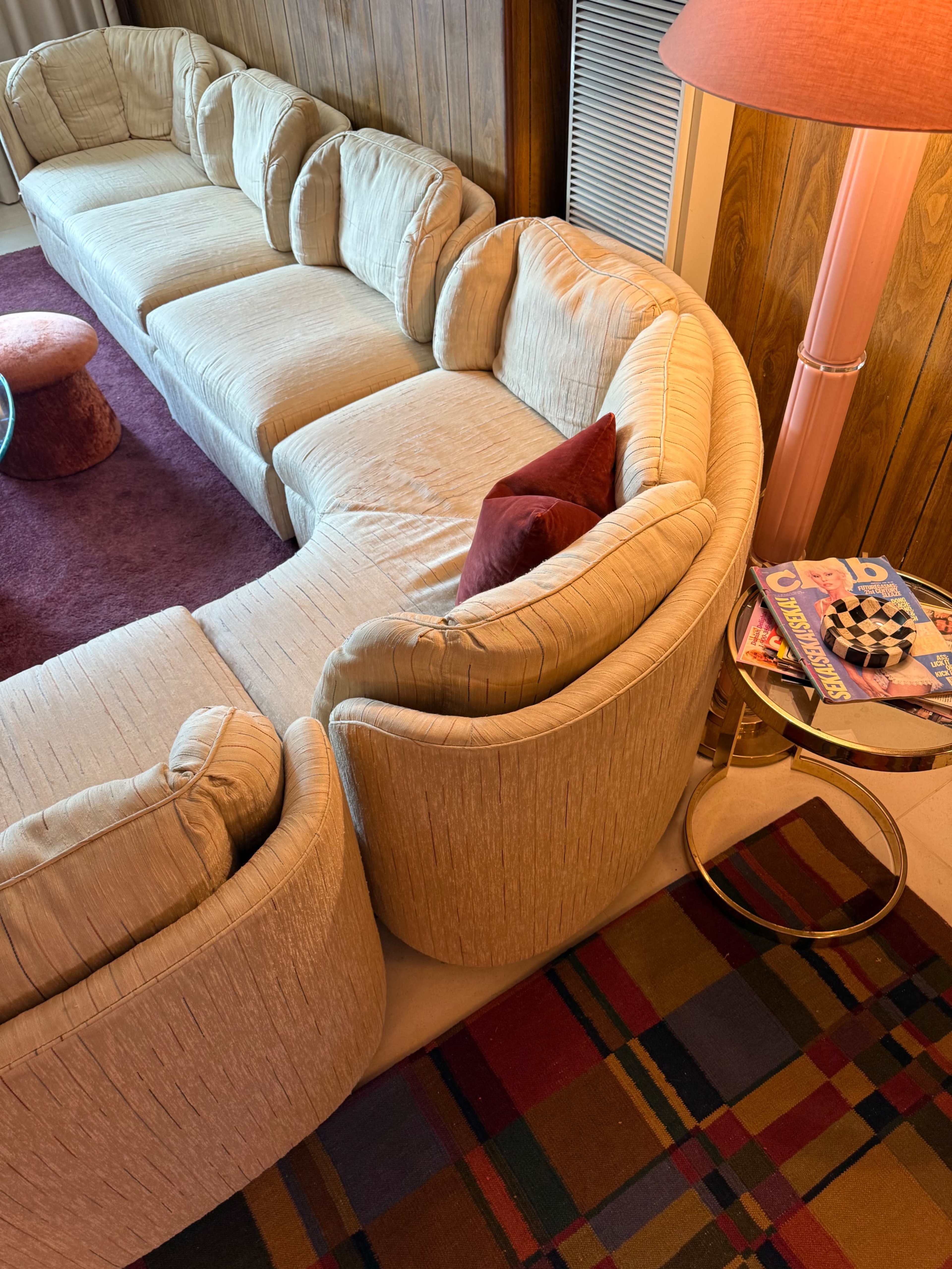 The image shows a curved, beige sectional sofa with a red cushion placed on it, positioned in a room with a patterned rug and a side table holding magazines.
