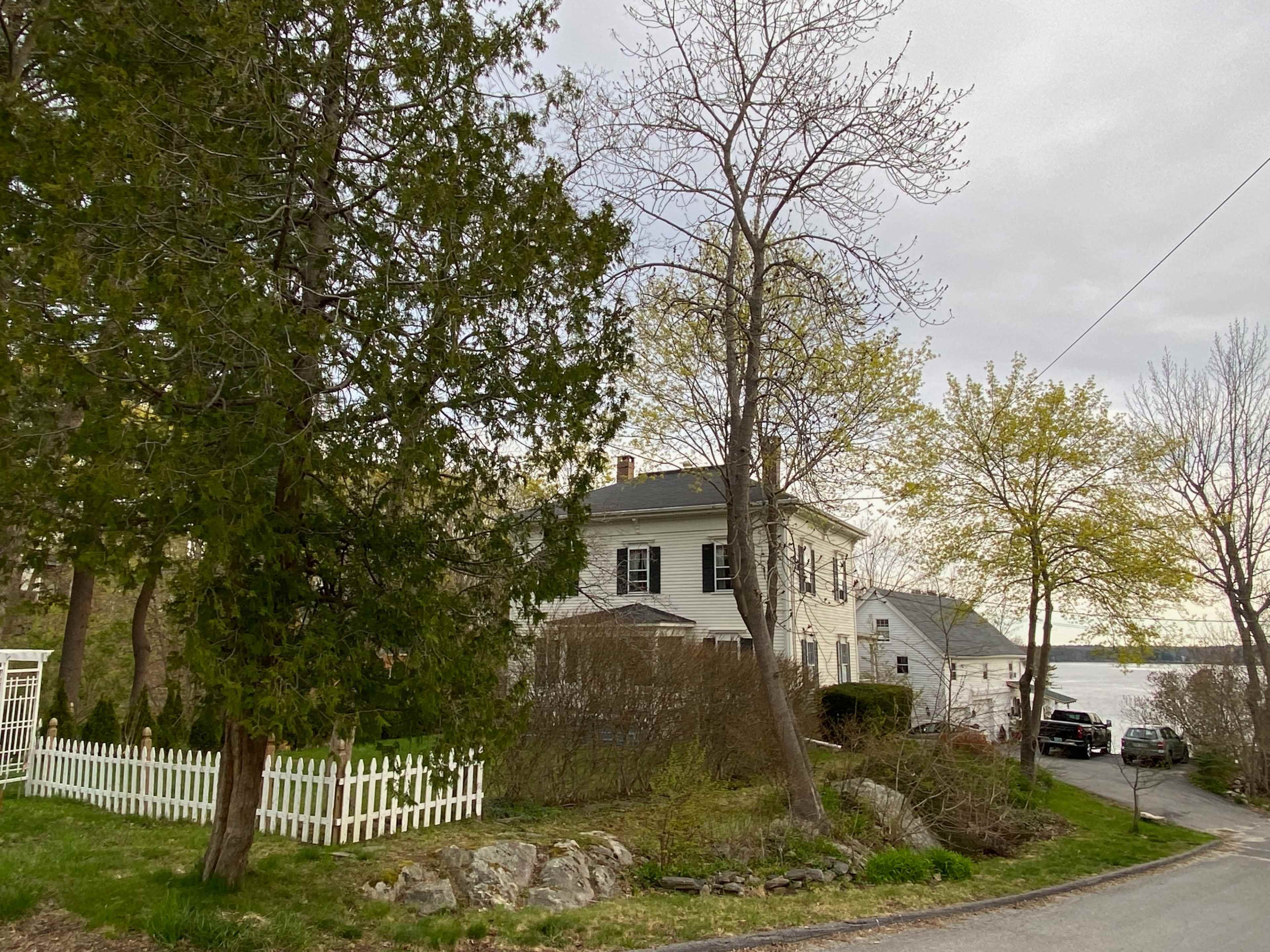 A house with a white picket fence is located near the water, surrounded by trees and low shrubs.