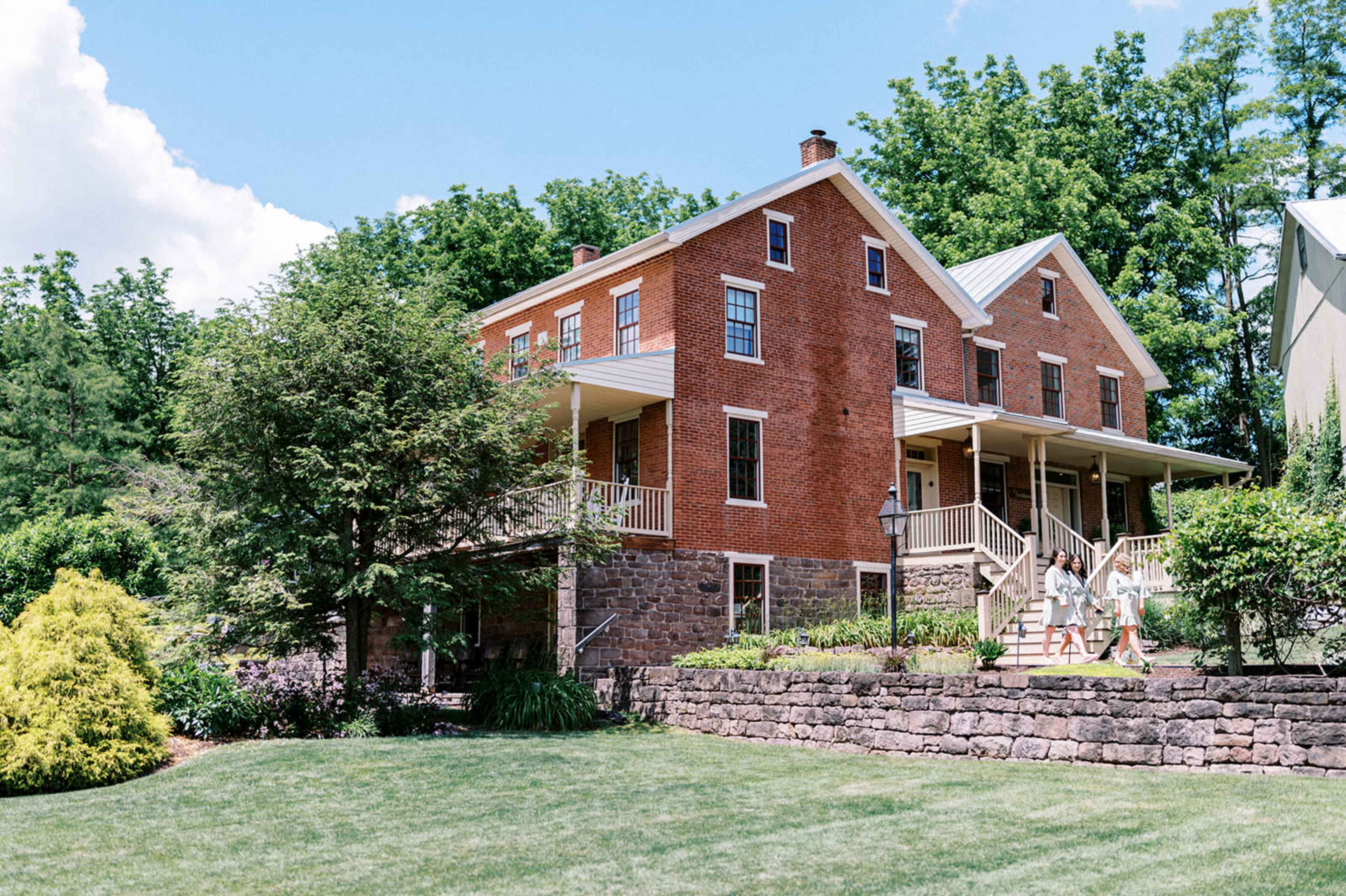 A two-story red brick house with a metal roof is surrounded by lush green trees and gardens.