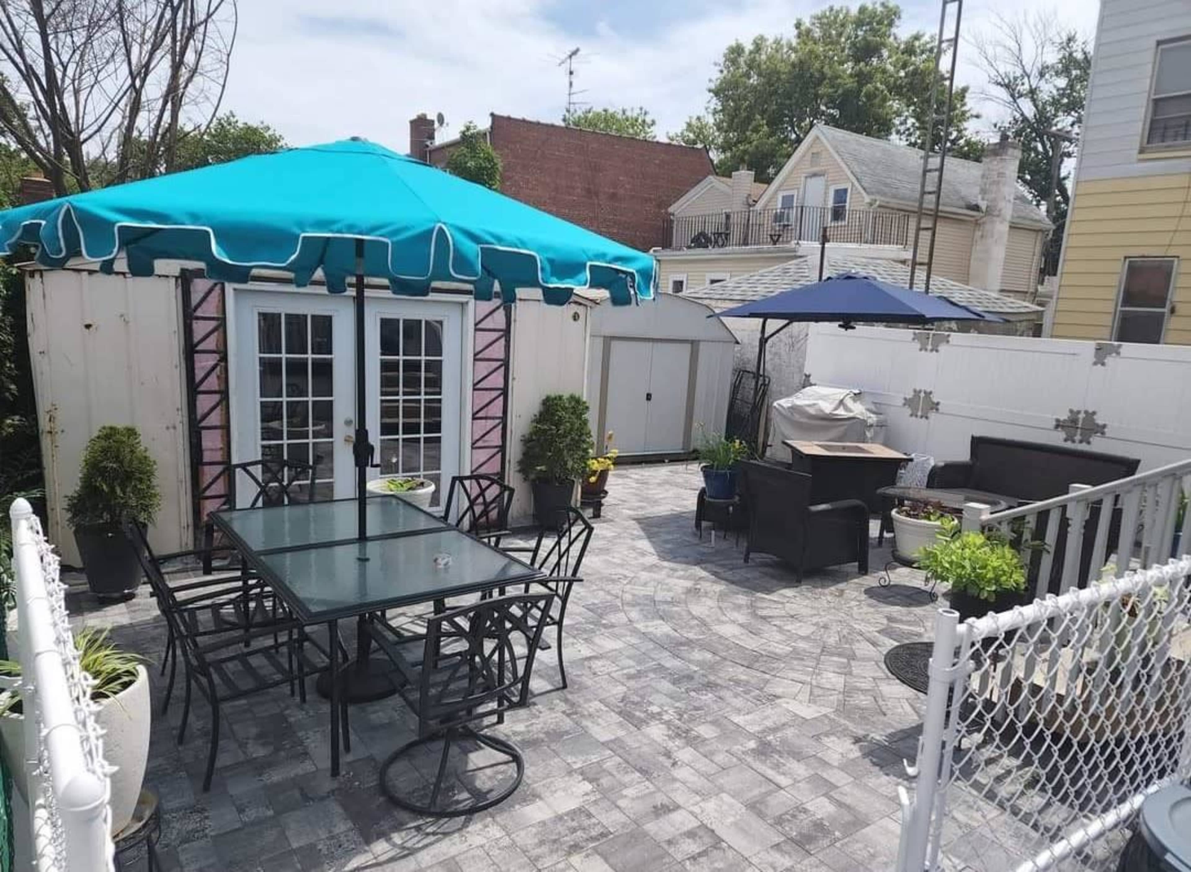 A fenced backyard patio with a large umbrella over a dining table, surrounded by potted plants and outdoor furniture.