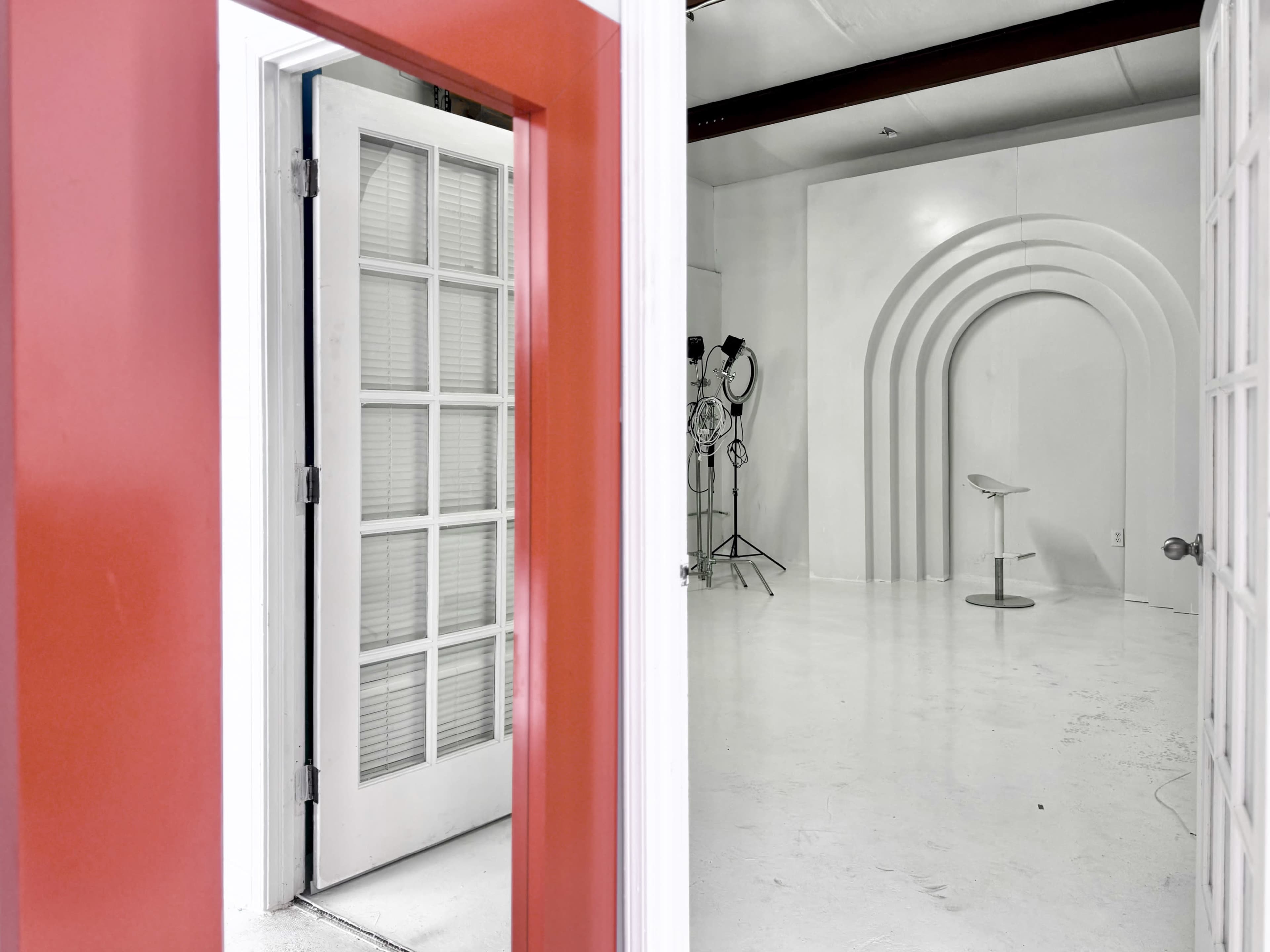 The image shows a doorway leading to a white studio space with a curved backdrop and a stool positioned in the center.
