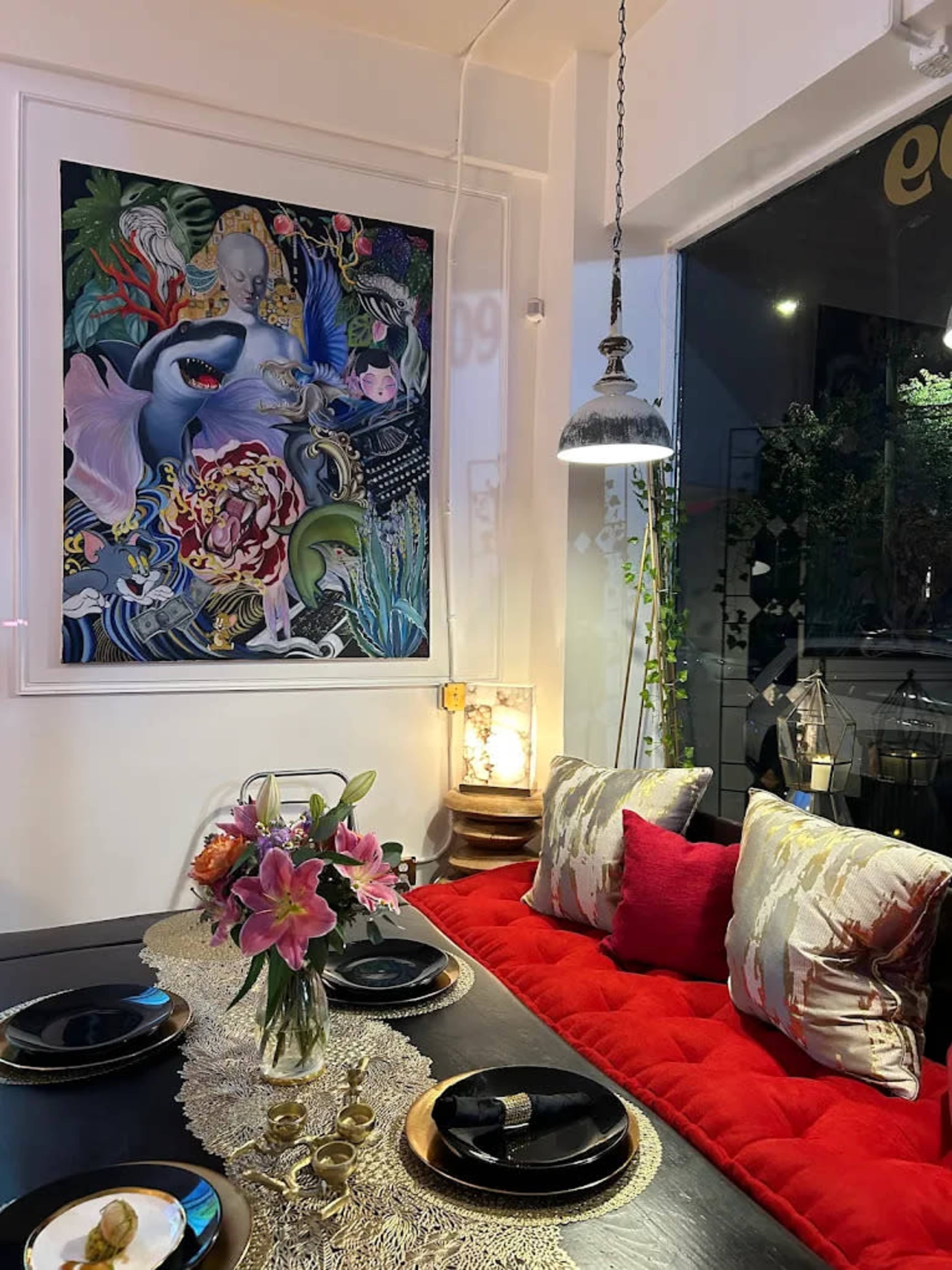 The image shows a cozy dining area featuring a large, colorful artwork on the wall, a table set with black plates and a floral centerpiece, and plush seating with decorative cushions.