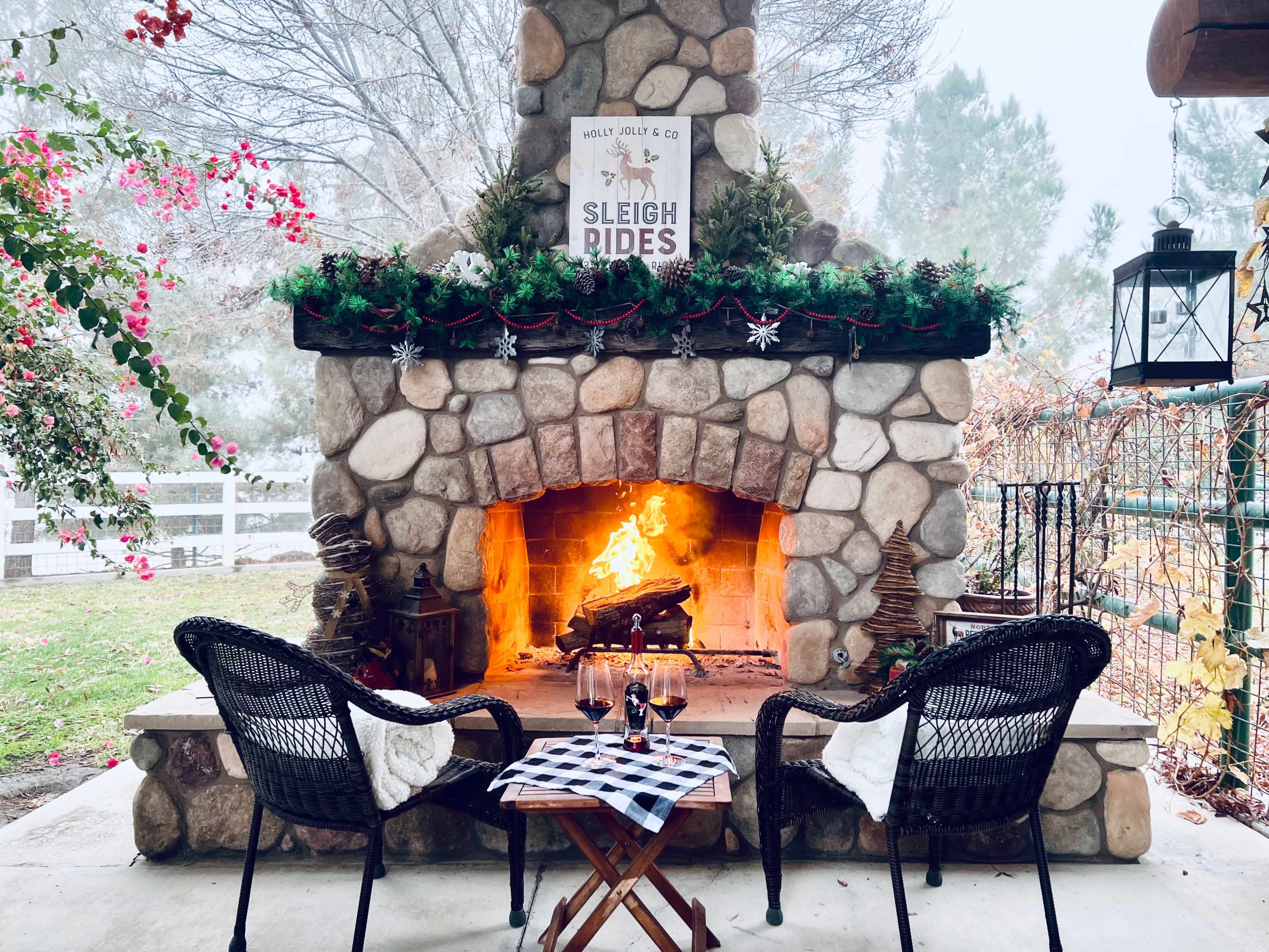 A stone fireplace with a roaring fire is flanked by two wicker chairs, and a sign reading "Sleigh Rides" is displayed above, surrounded by greenery and seasonal decorations.