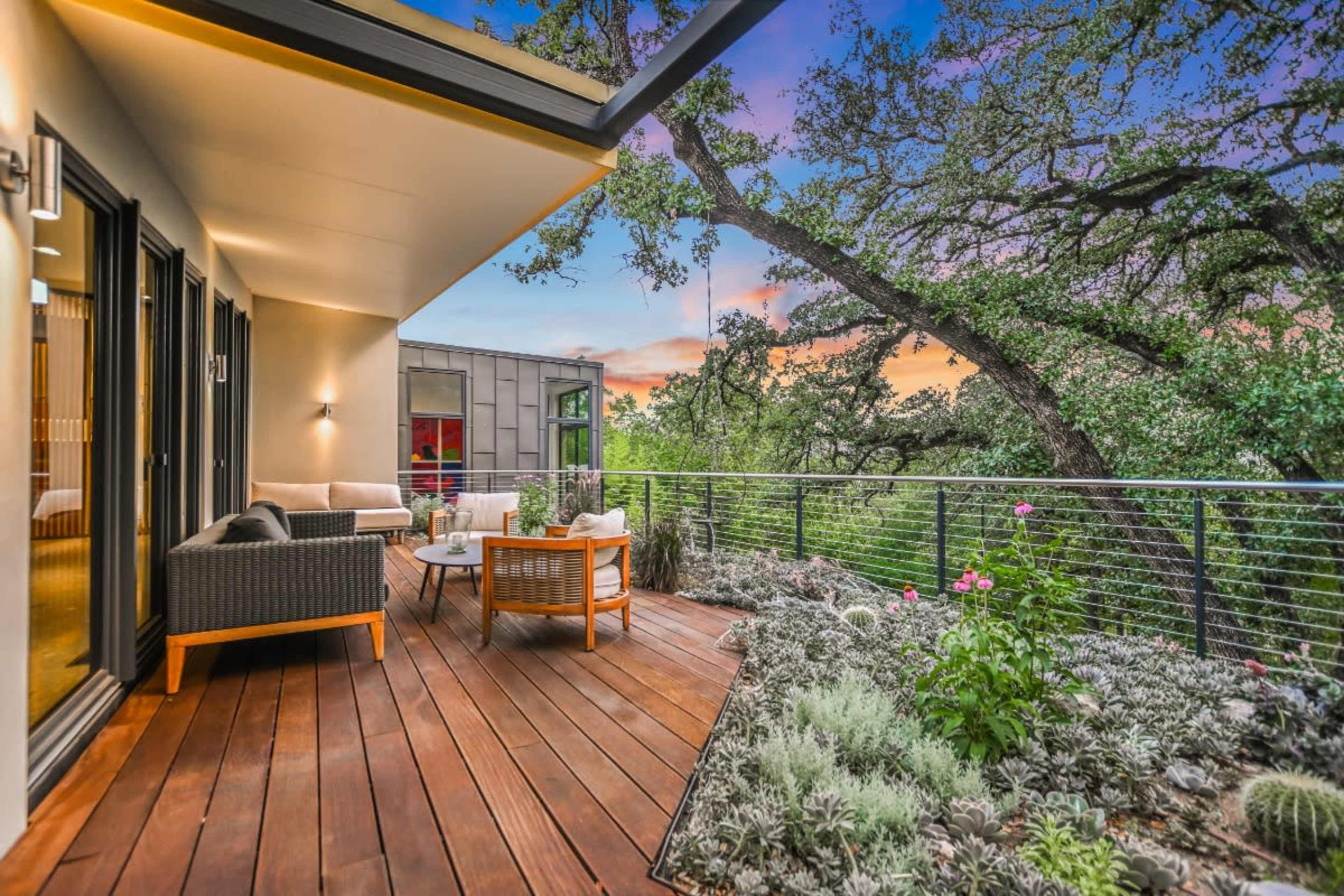A modern wooden deck features two seating areas surrounded by lush greenery and a sunset sky in the background.