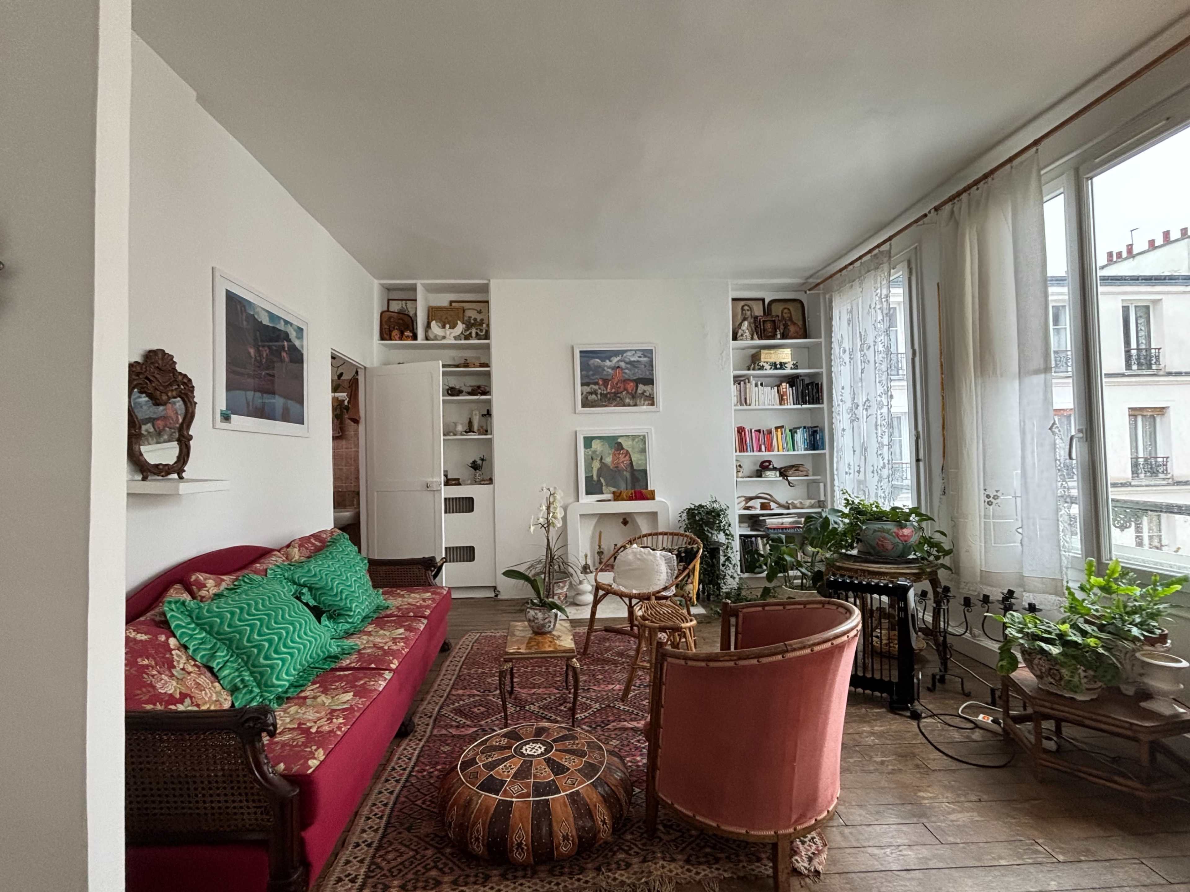 The image shows a bright living room with a red couch, a round coffee table, shelves with decor, and several indoor plants by large windows.