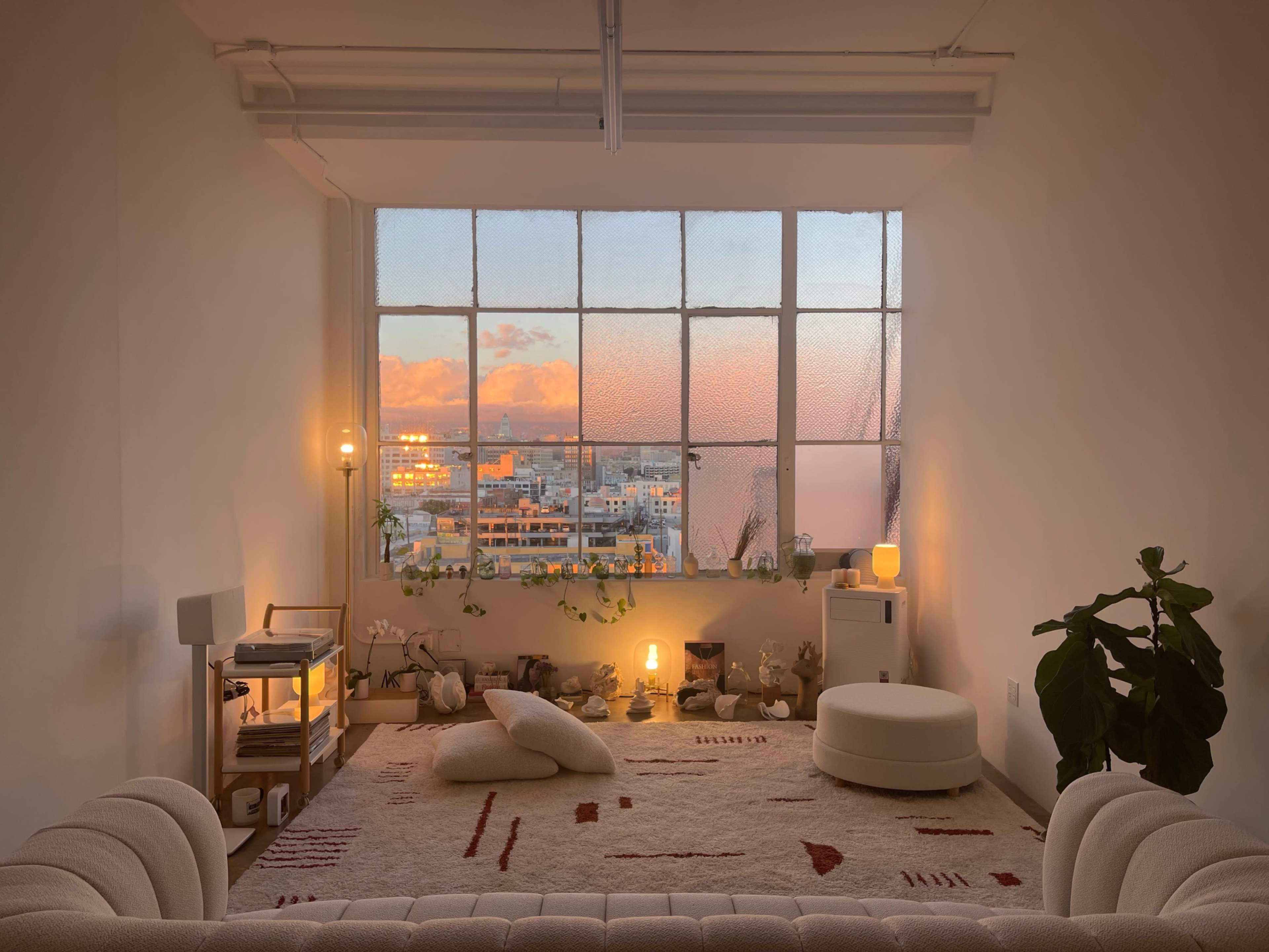 The image shows a cozy, well-lit room with a large window revealing a sunset view, featuring a white couch, a decorative rug, and various plants.