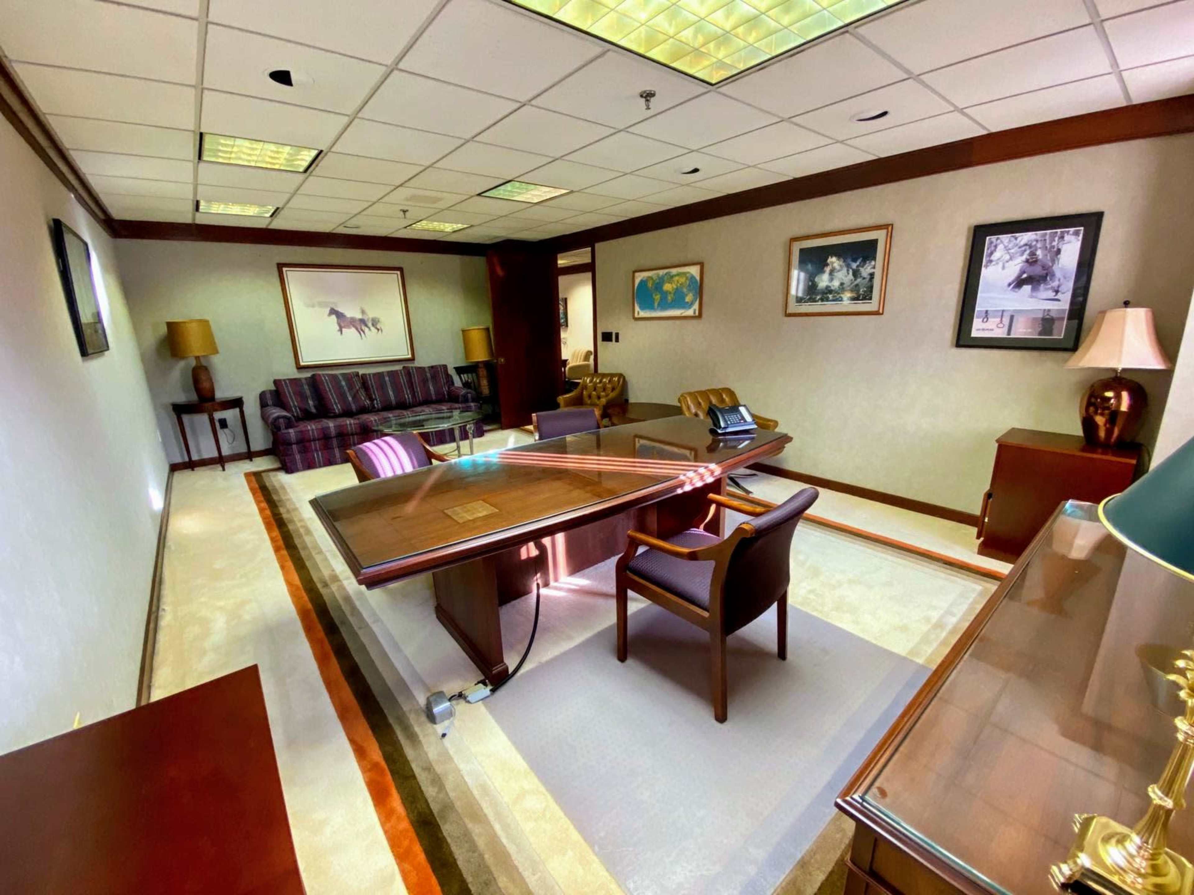 The image depicts a spacious office featuring a large wooden desk, leather chairs, a sofa, and framed artwork on the walls.