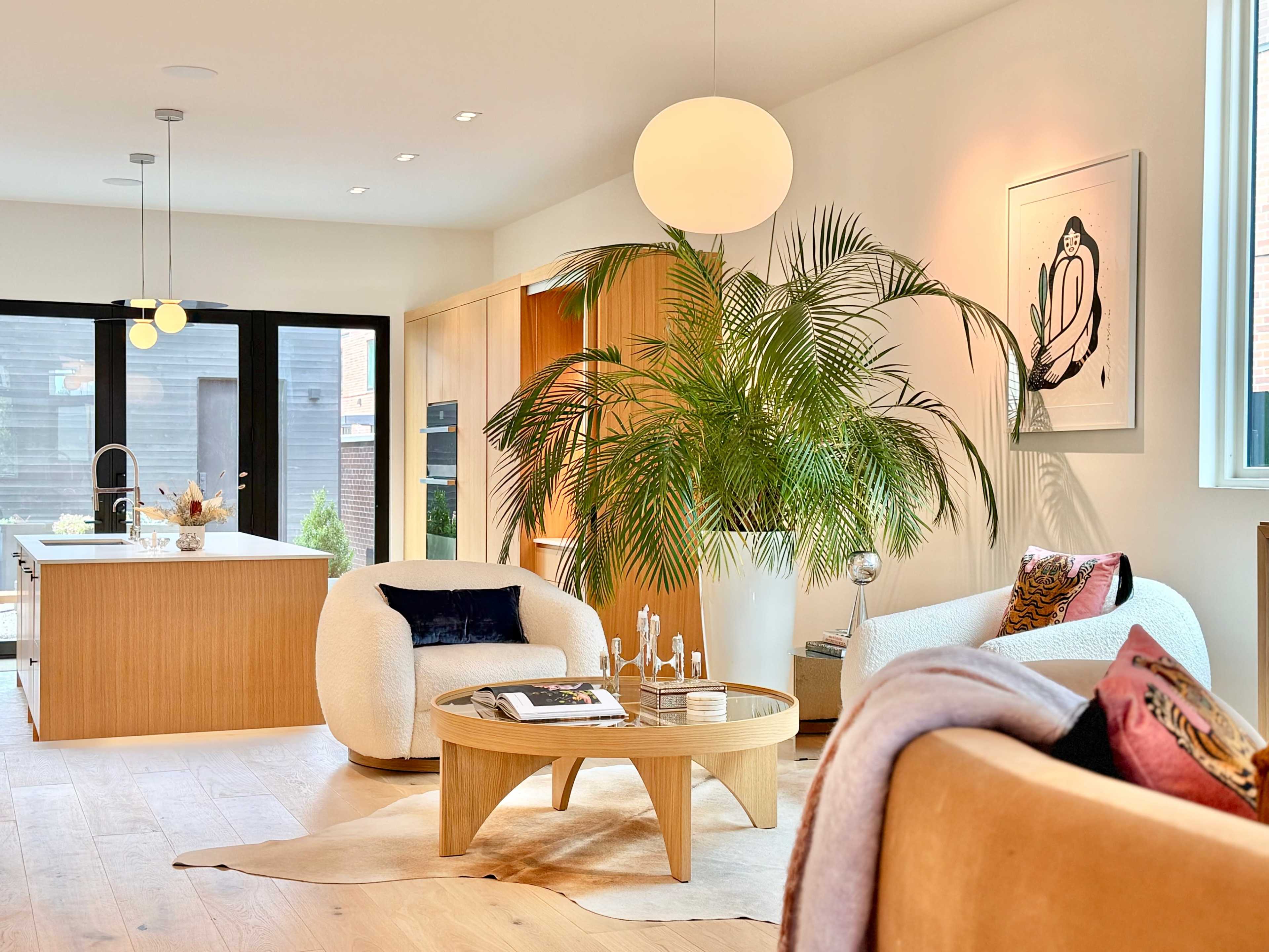 The image shows a modern living room featuring two soft chairs, a round coffee table, and a large potted plant, with a kitchen visible in the background.