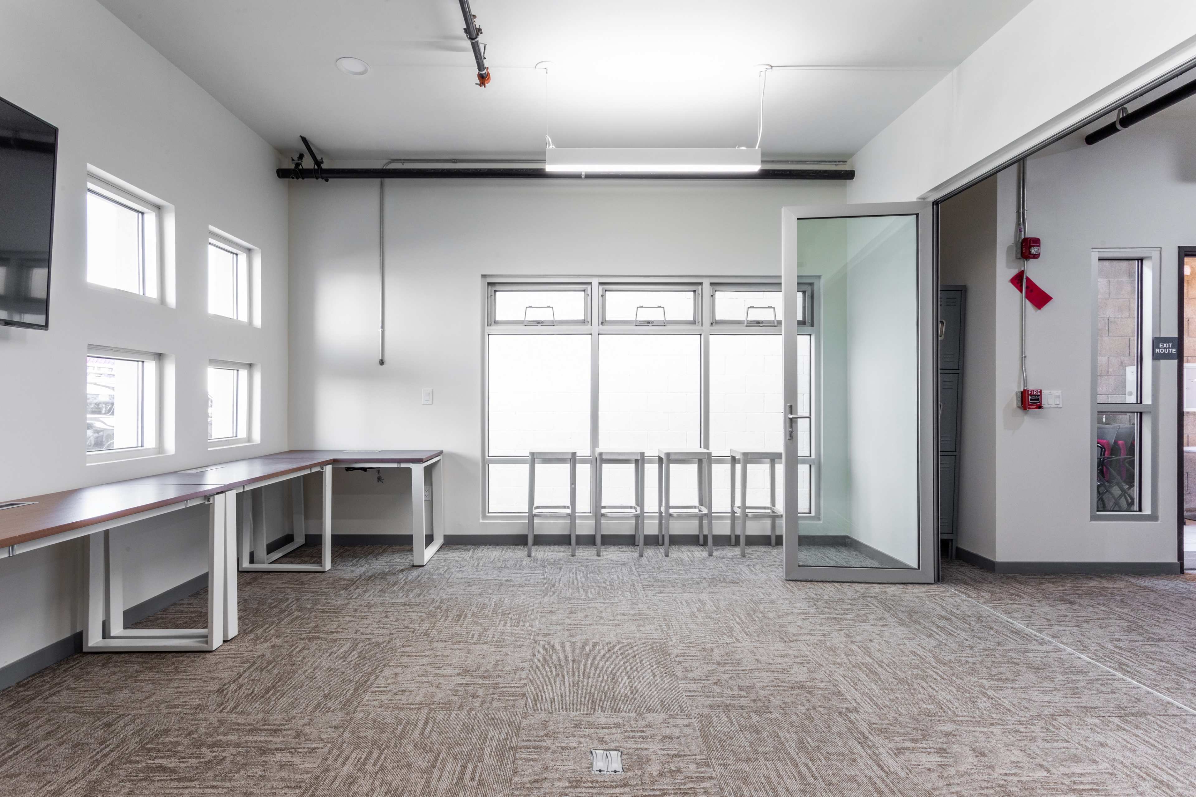 A spacious, empty room with a long table on one side and a set of metal stools by a window.