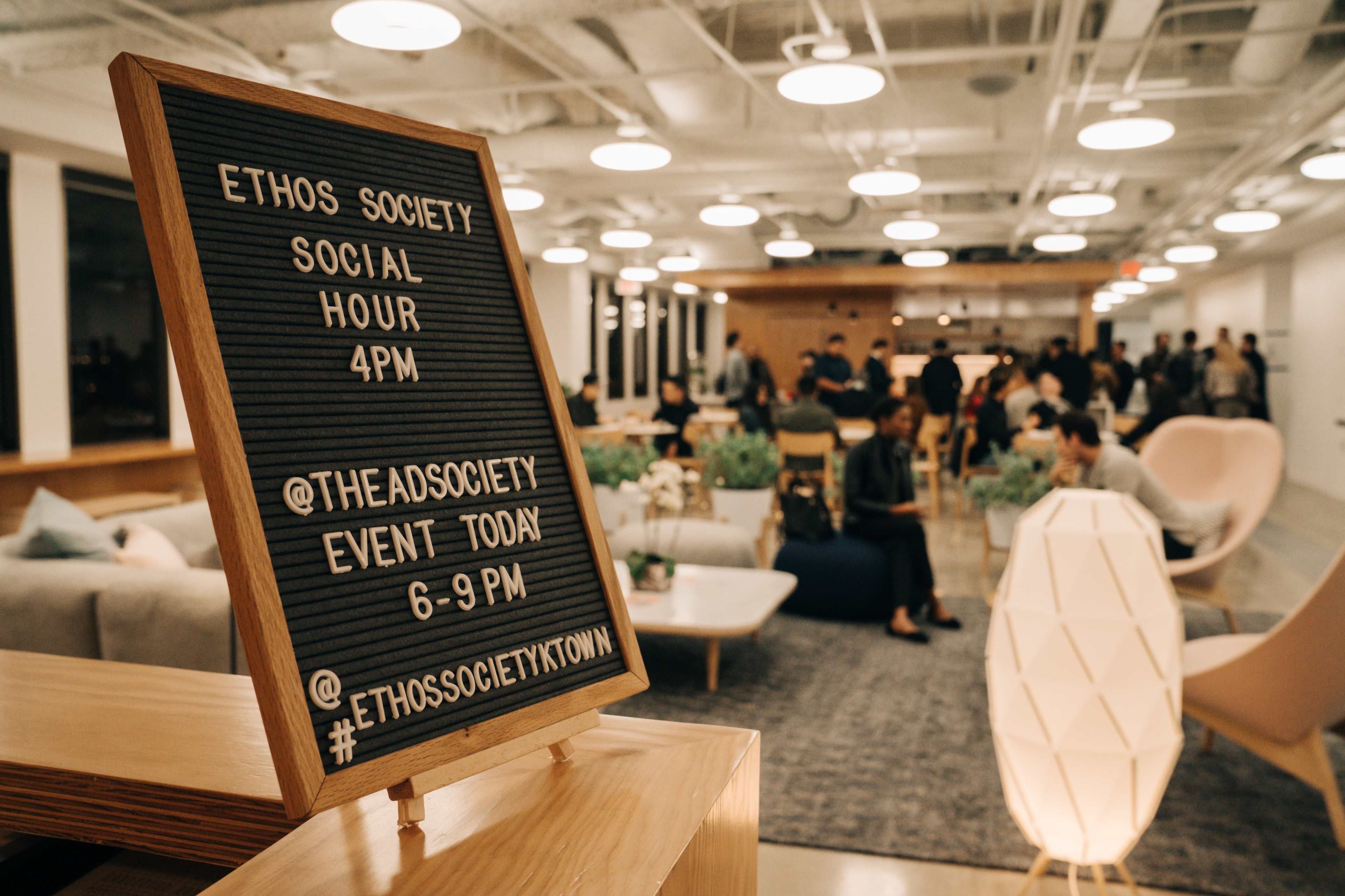A sign advertising the Ethos Society social hour and event is displayed in a modern indoor space filled with attendees.