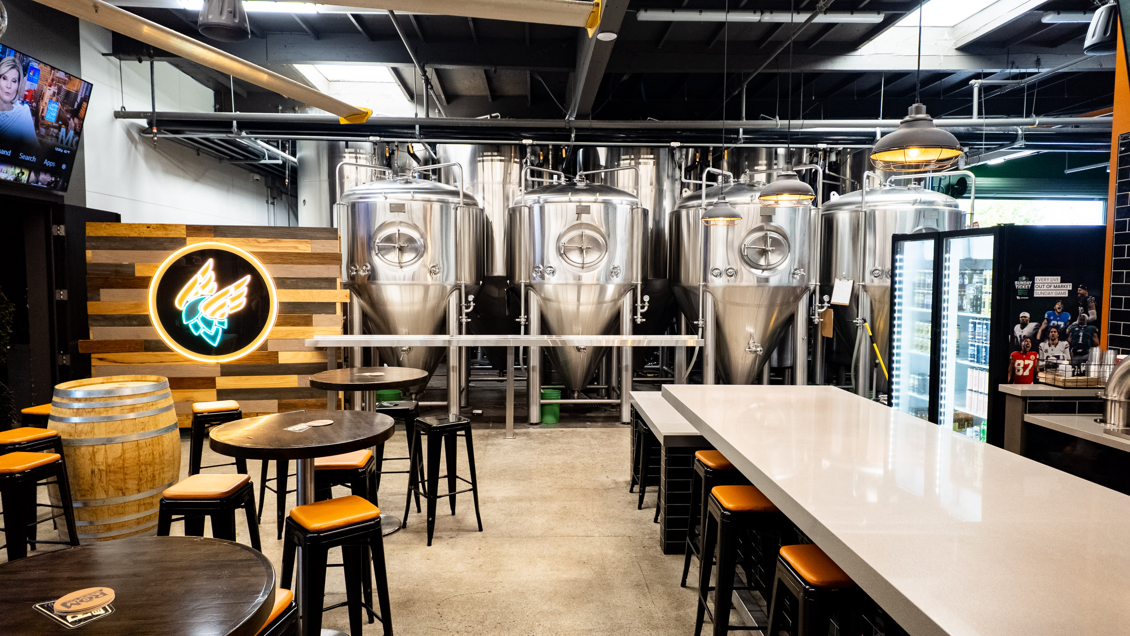 The image shows a modern brewery interior featuring stainless steel fermentation tanks, a bar area with tall tables and chairs, and a neon sign on the wall.