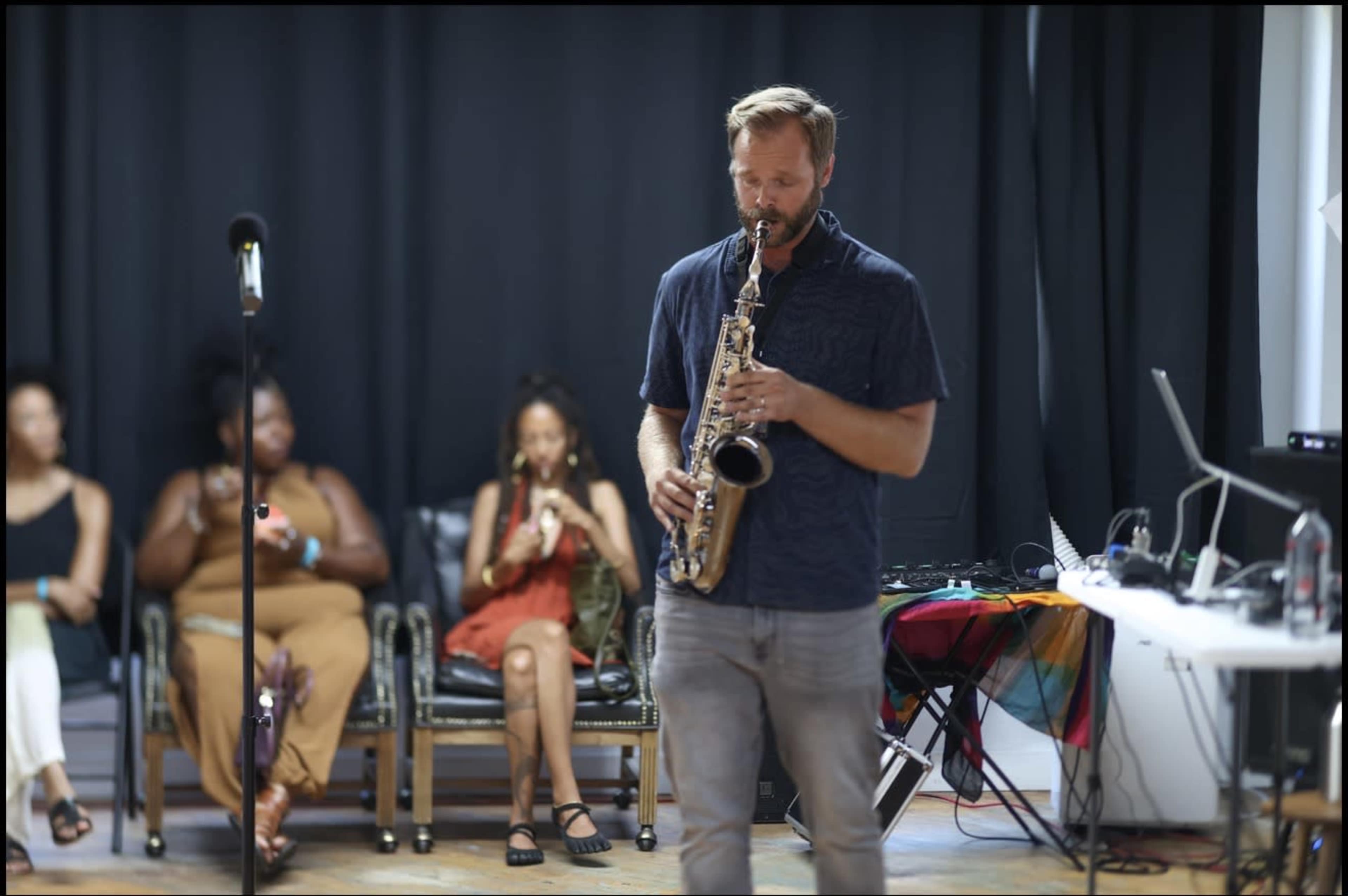 A musician plays a saxophone while an audience sits in chairs behind him.