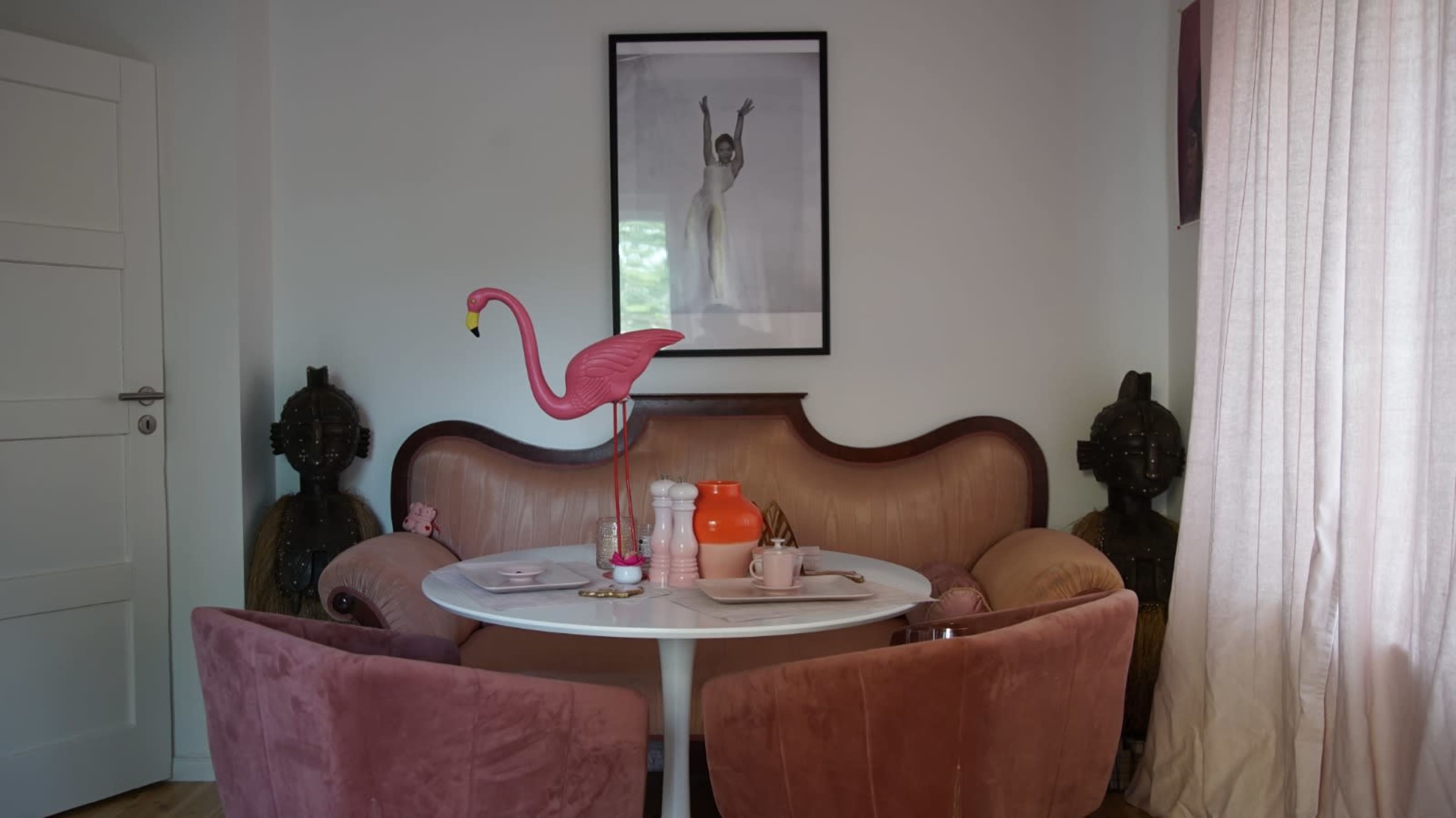 A round table sits in a cozy corner with pink chairs, a flamingo figure, and a decorative orange vase against a backdrop of a framed black and white photograph.