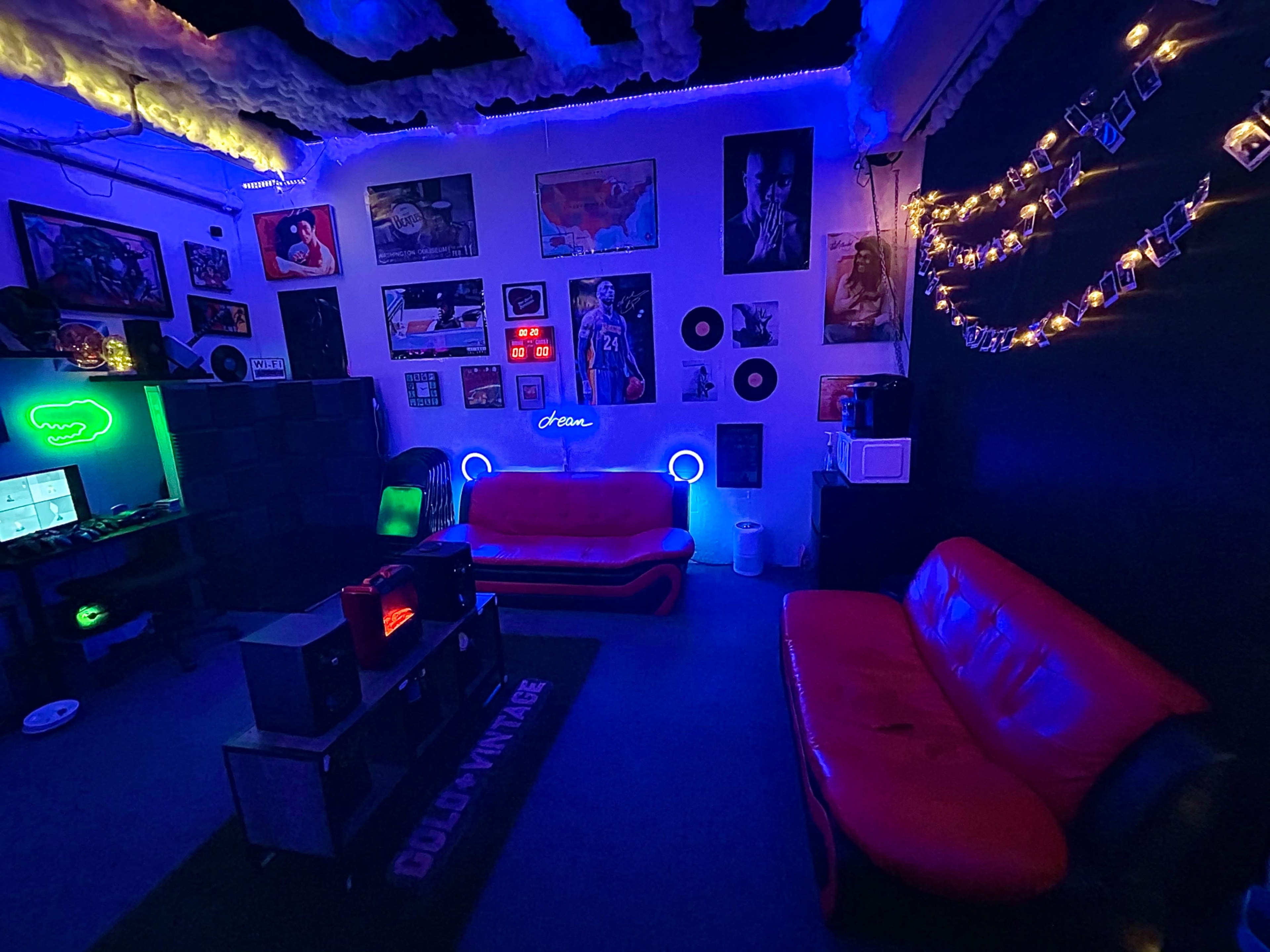 The image shows a dimly lit room with red couches, colorful lighting, and various posters and decorations on the walls.