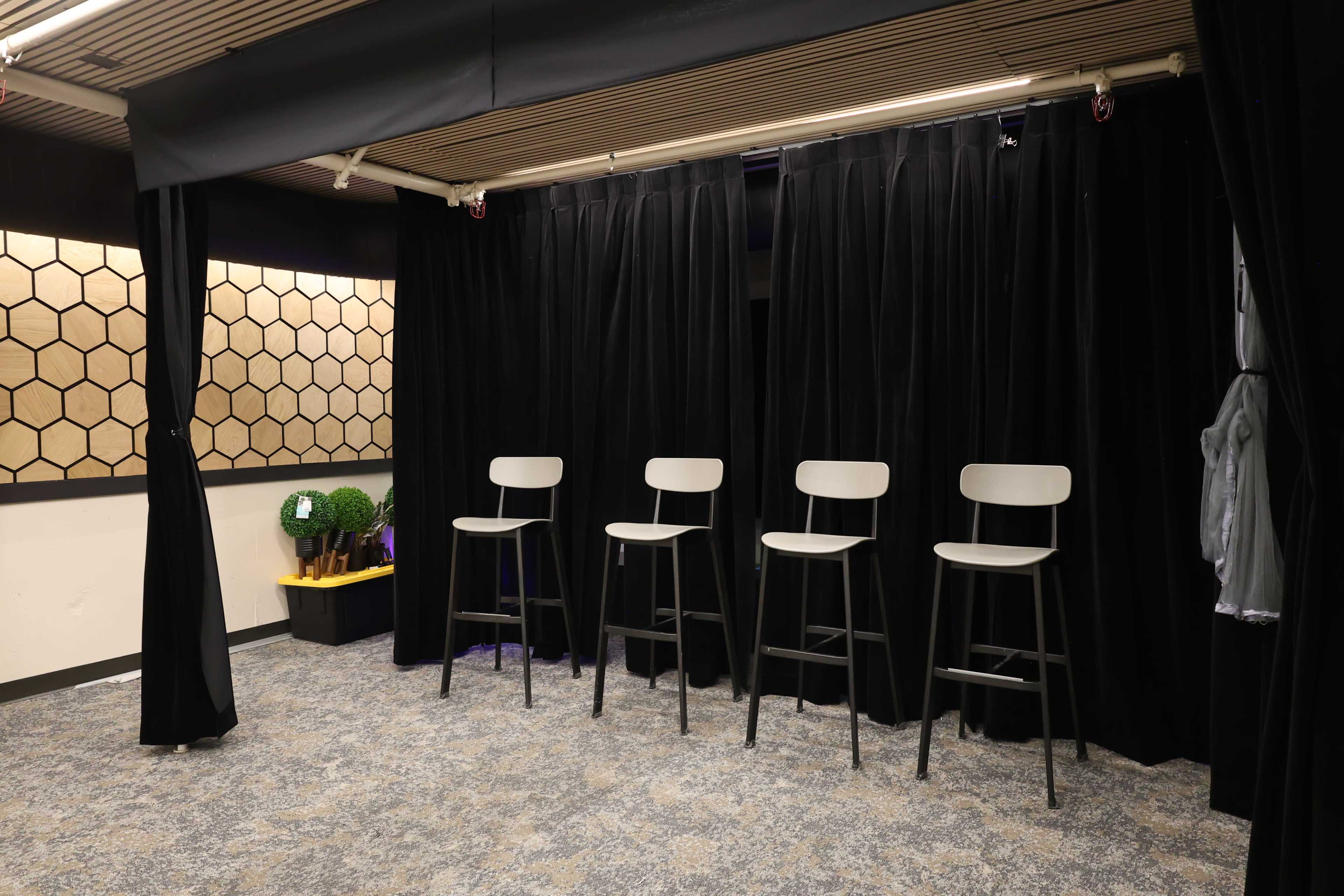 The image shows a small stage area with four high stools positioned in front of a black curtain, set against a backdrop of hexagonal panels.