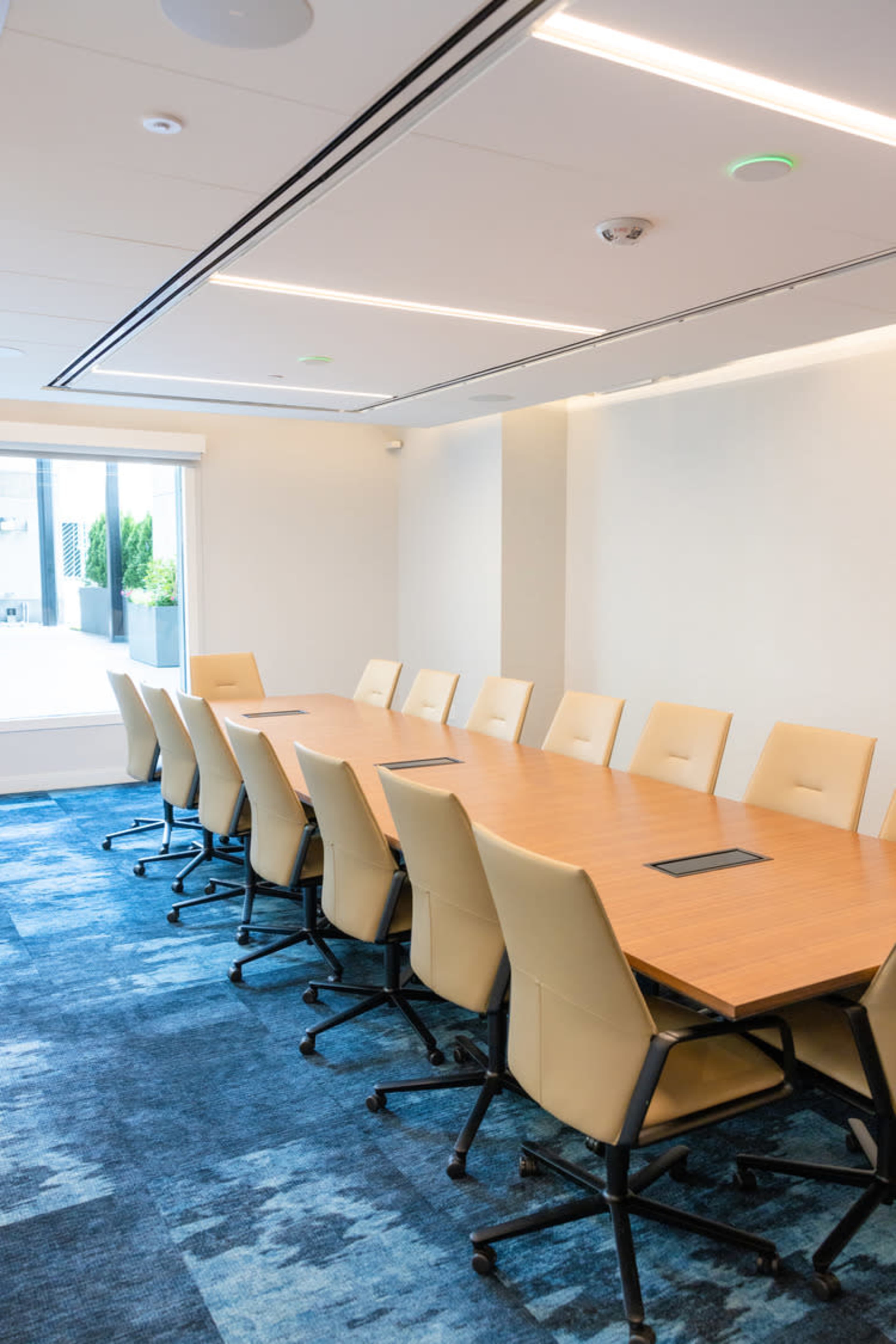 A spacious conference room with a long wooden table surrounded by beige chairs, featuring a large window that allows natural light to enter.