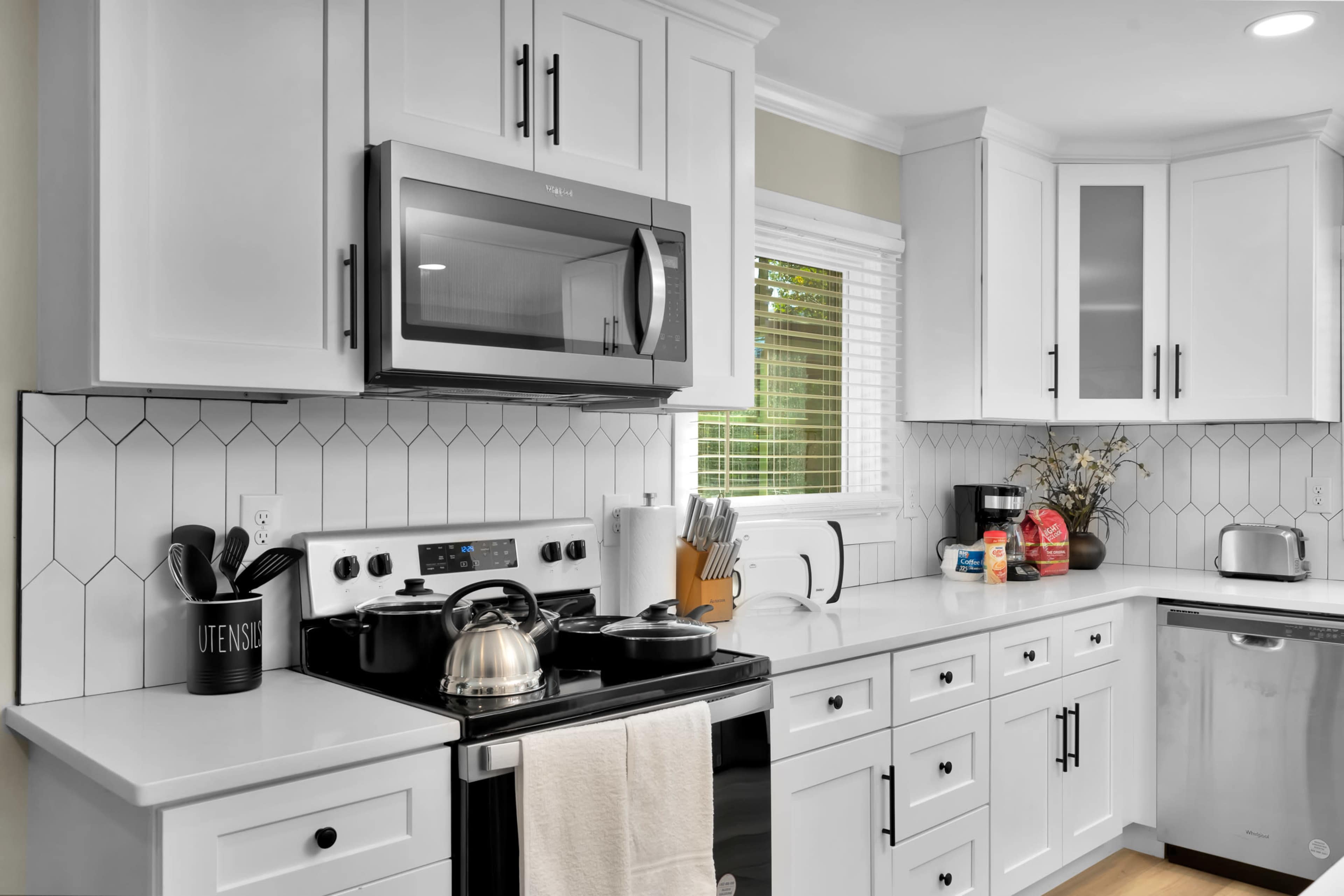 The kitchen features white cabinets, a stainless steel microwave, and a modern stovetop with a kettle, arranged alongside a tiled backsplash and ample countertop space.