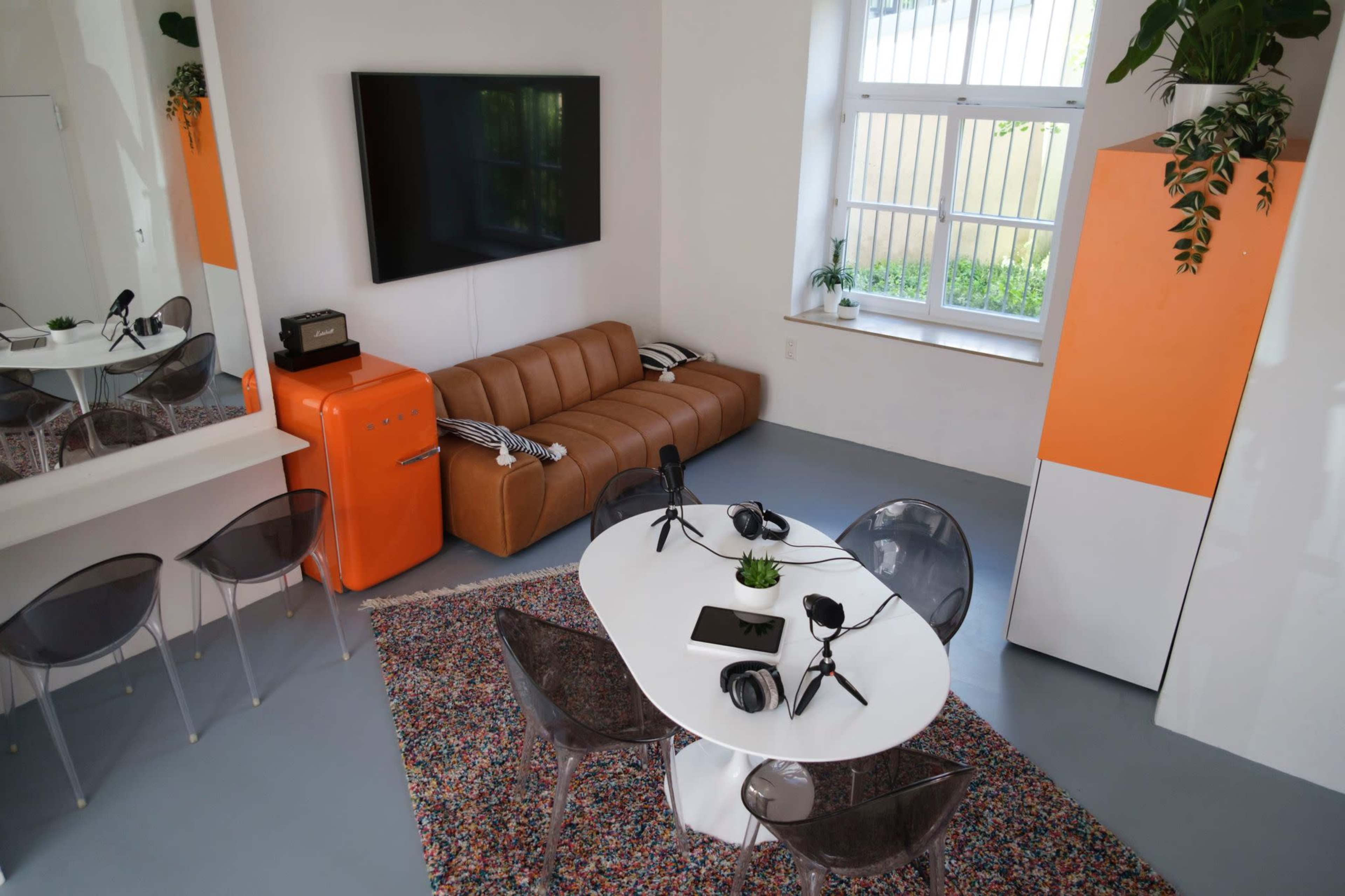 The image shows a modern, well-lit room featuring a brown leather sofa, a white dining table with microphones, and an orange refrigerator, all complemented by a colorful rug and indoor plants.