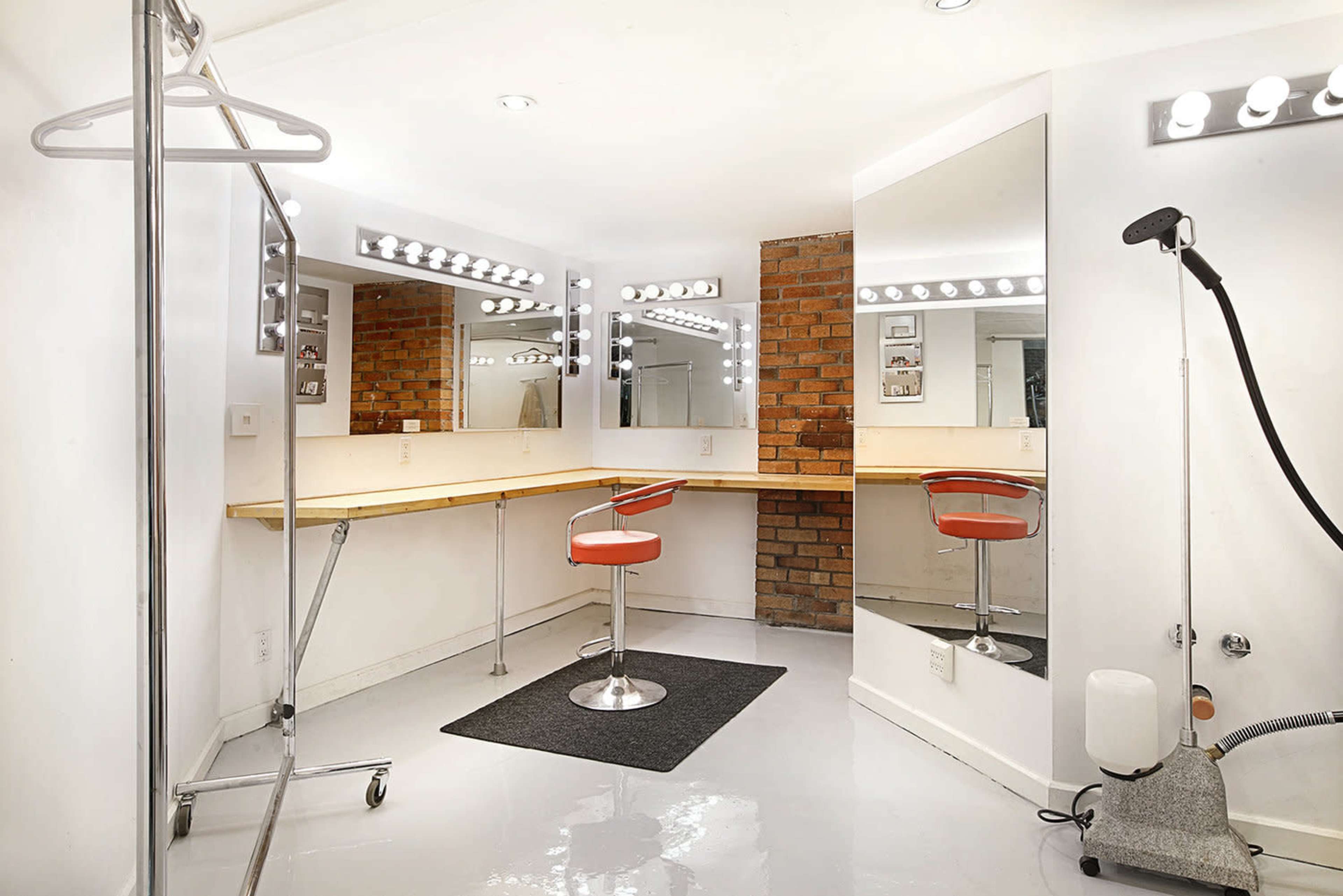 The image shows a well-lit dressing room with a large mirror, a wooden counter, an orange swivel chair, and a clothing rack.