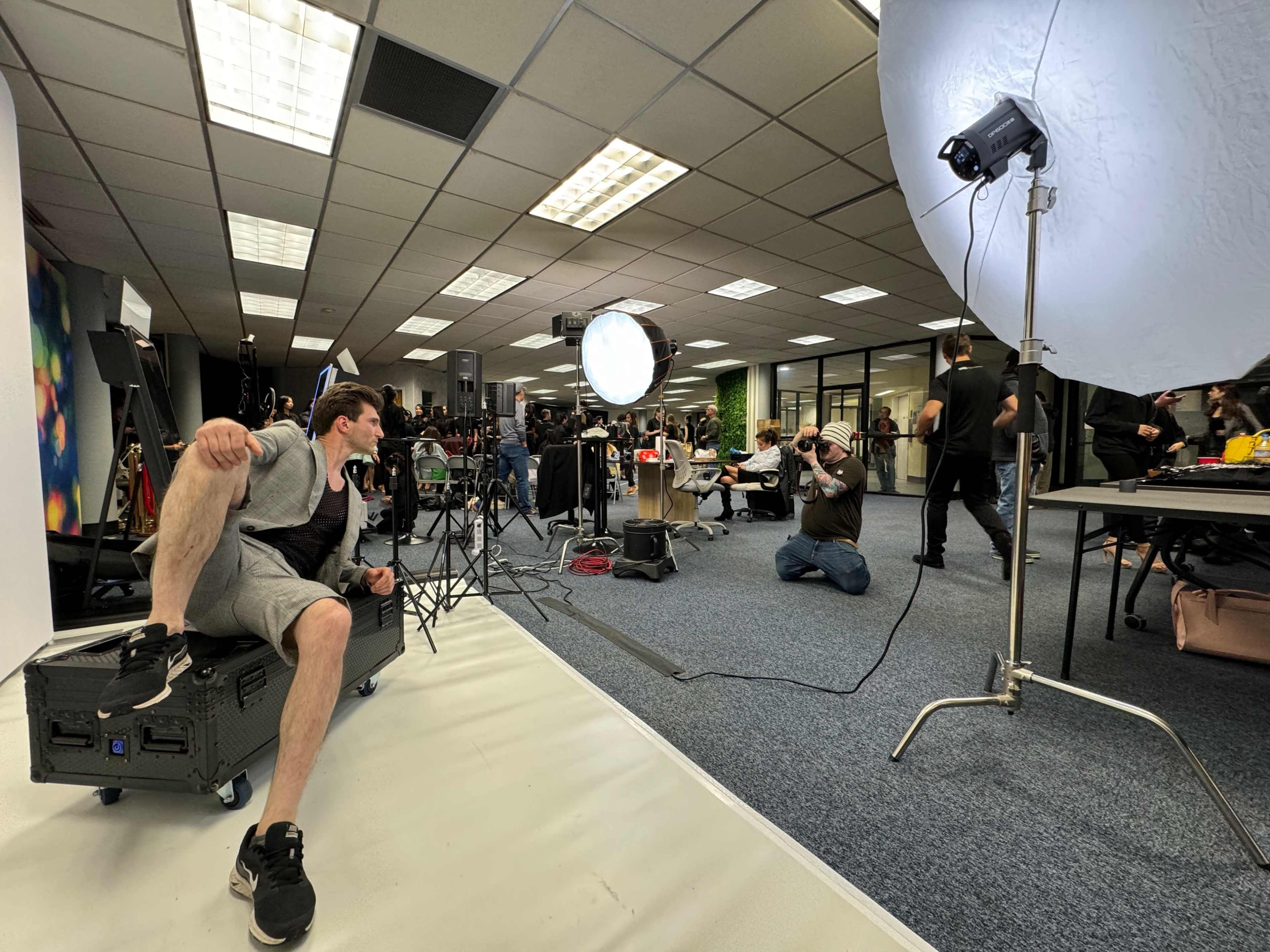 A photographer captures images of a seated model in a studio filled with various equipment and people.