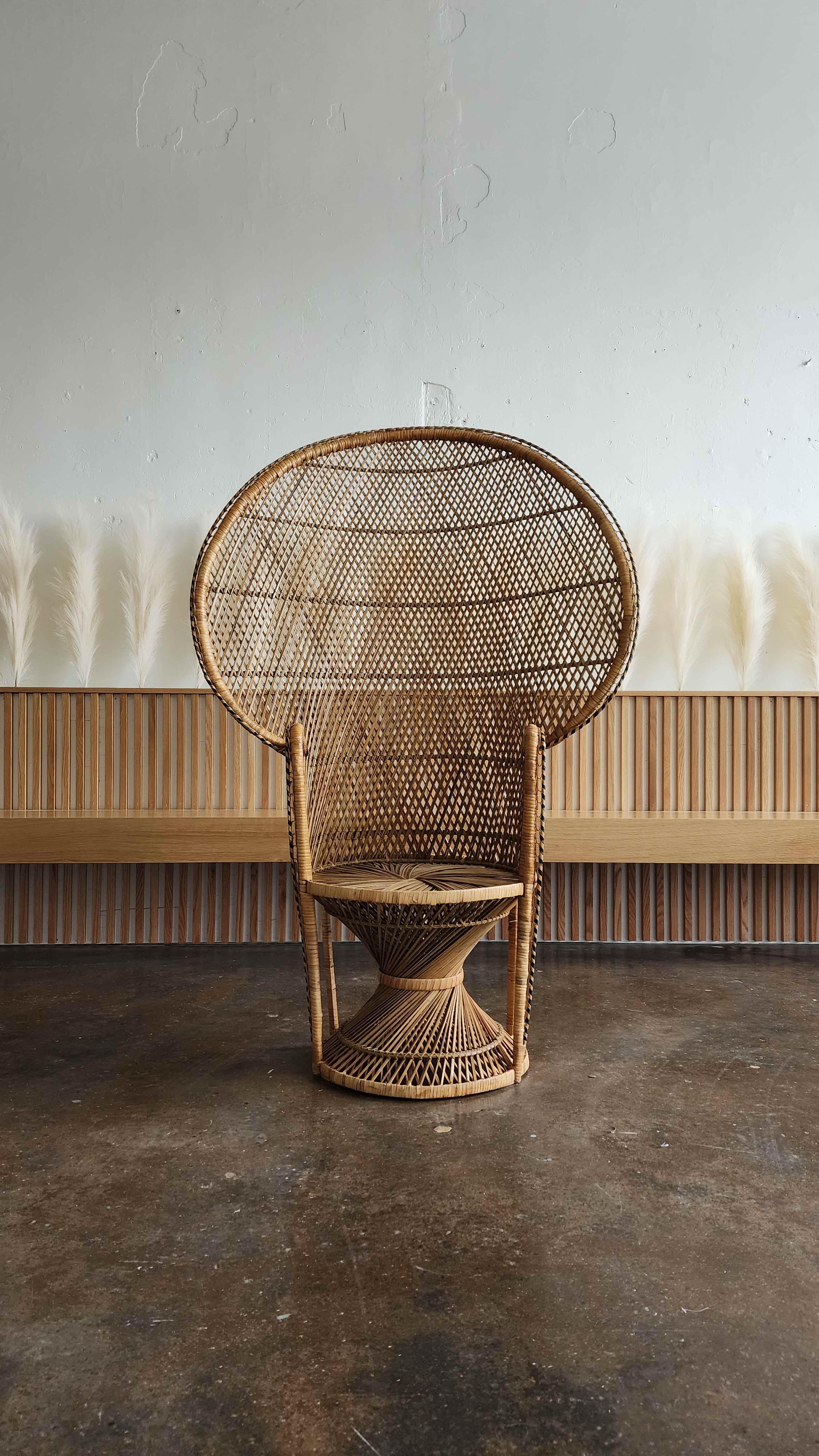 A large, intricately woven rattan chair is positioned against a plain wall, with a wooden bench and decorative pampas grass in the background.