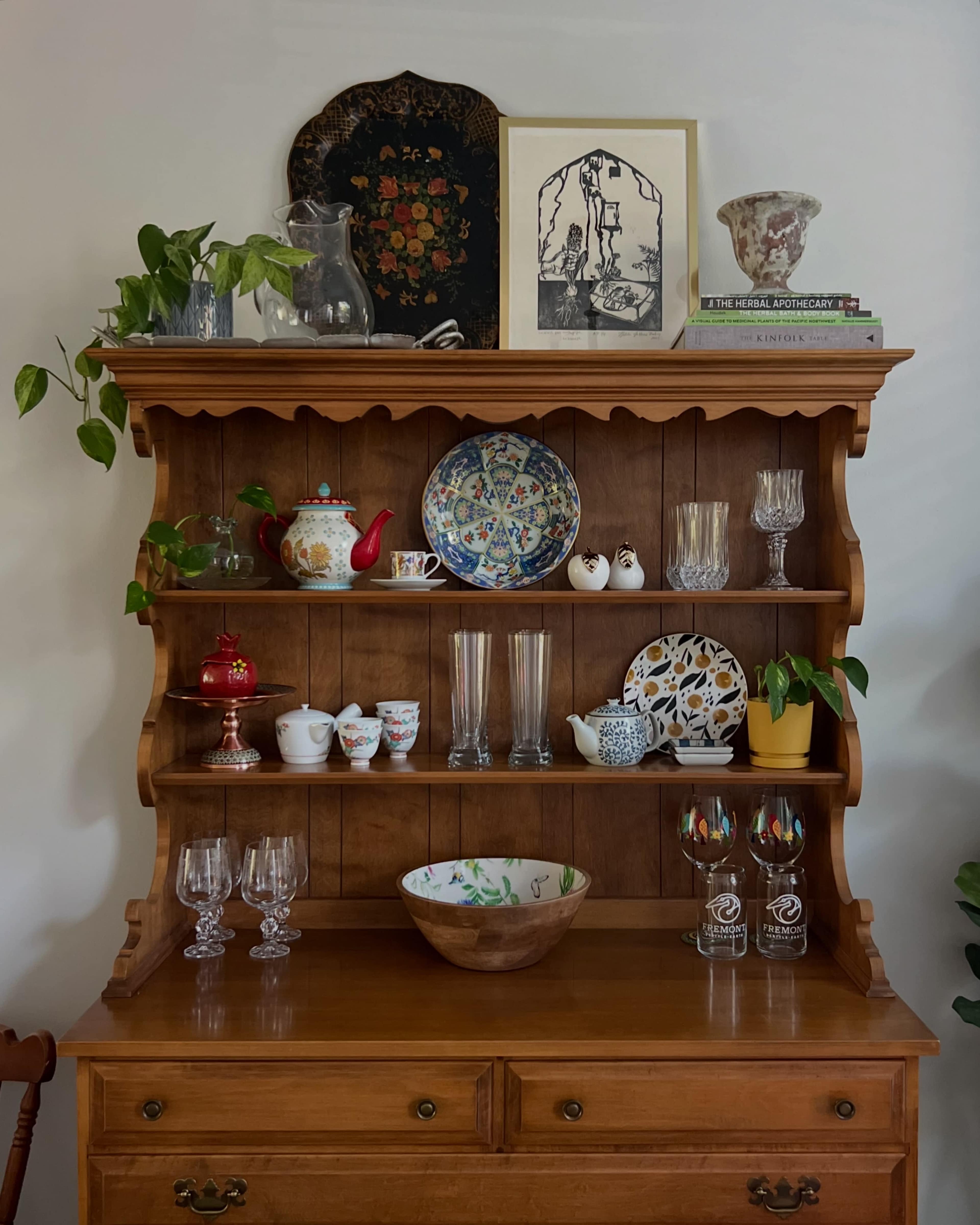 A wooden hutch displays a variety of decorative dishes, glasses, and plants, with a framed artwork above the shelf.