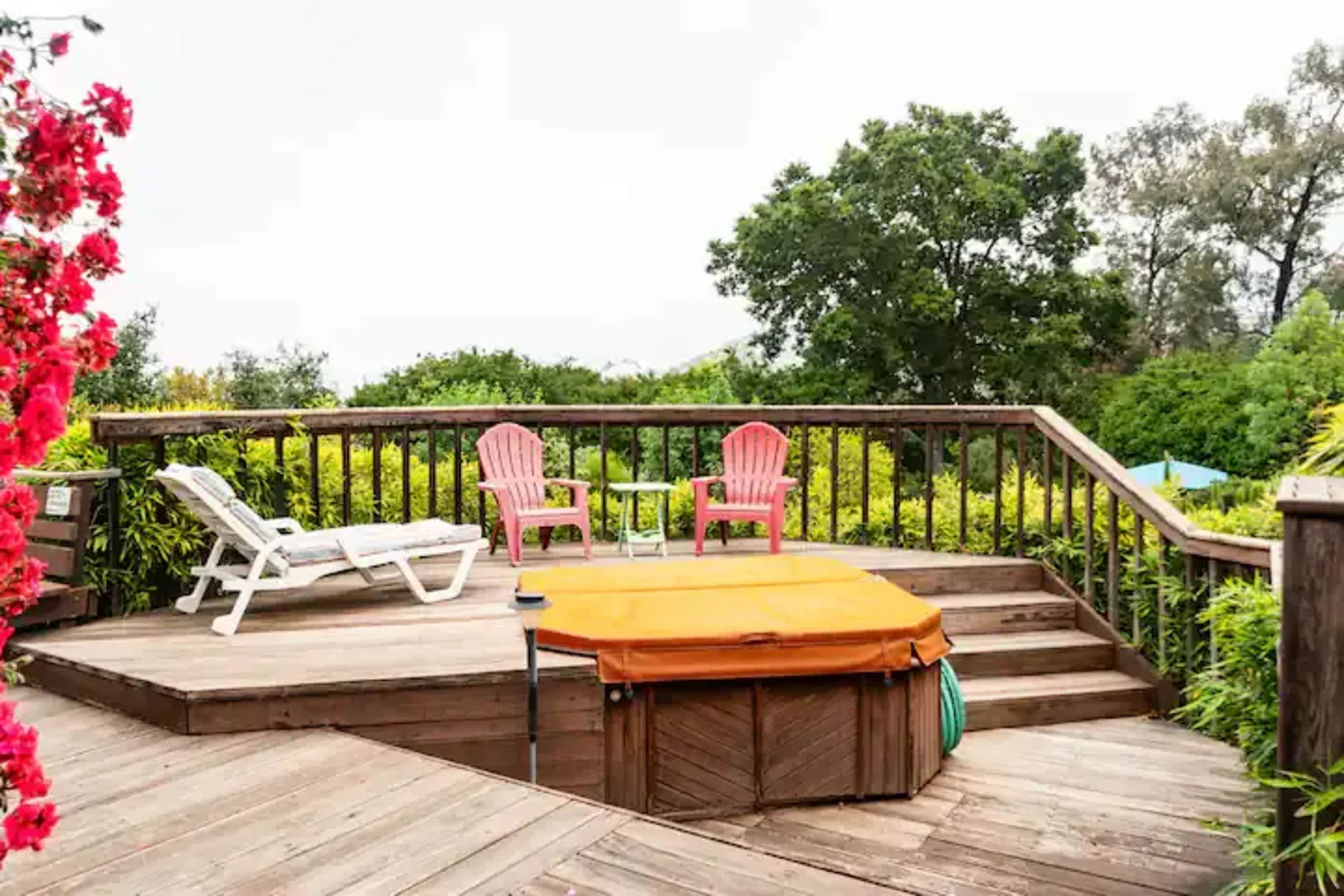 A wooden deck features a hot tub and two pink chairs, surrounded by lush greenery and colorful flowers.