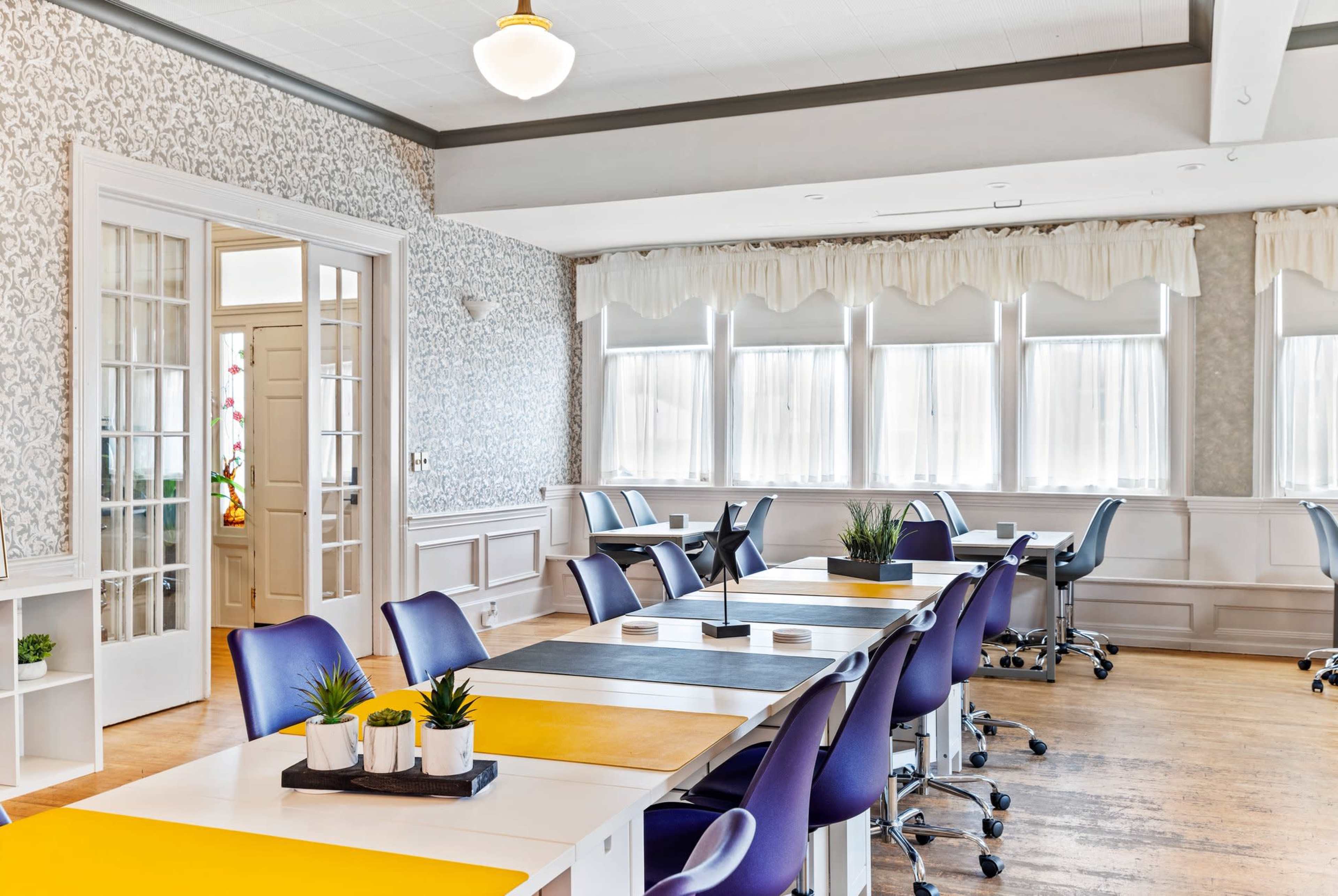 A bright, modern office space with multiple workstations featuring white tables, purple chairs, and large windows allowing ample natural light.