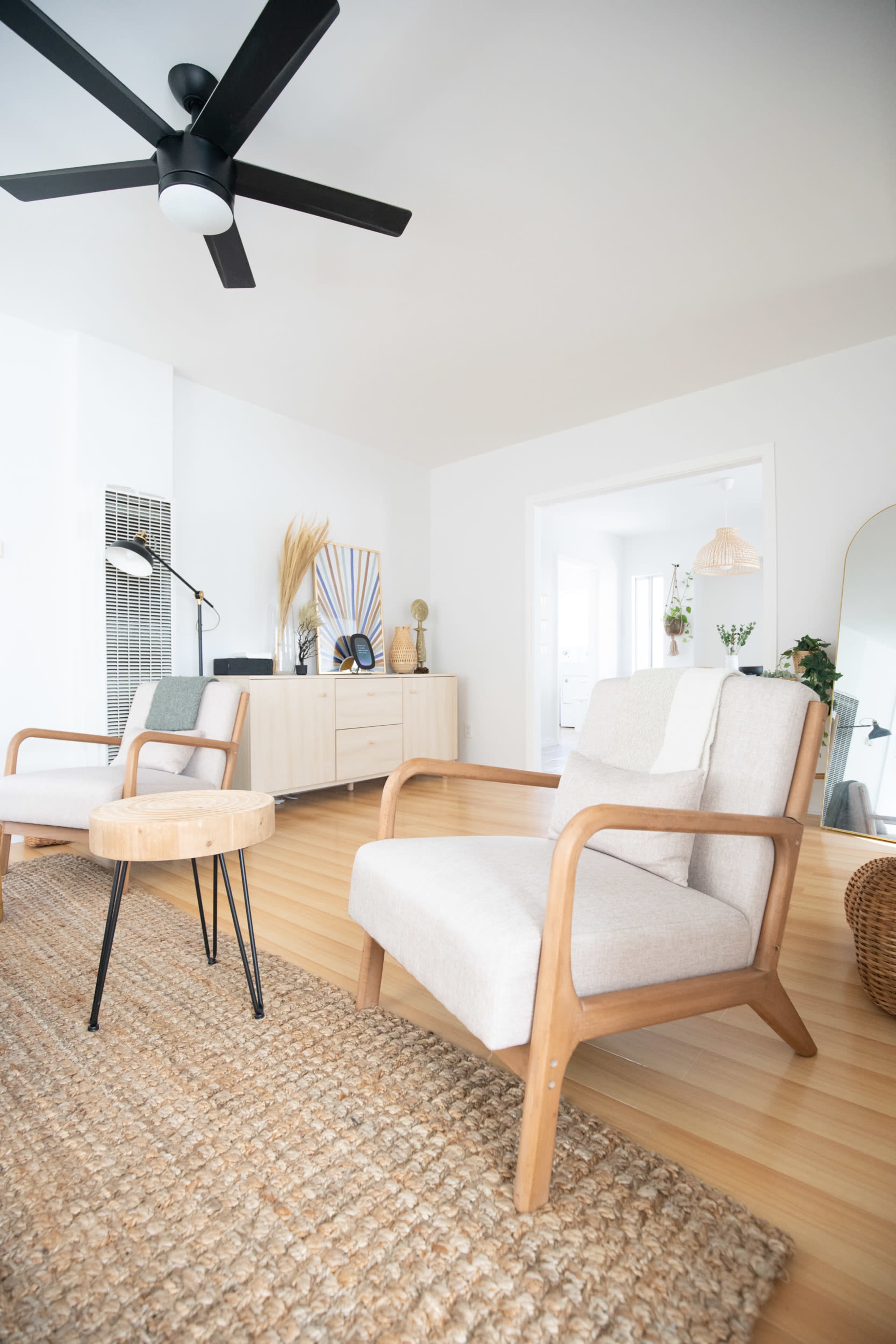 A bright living room with two wooden-framed chairs facing a small circular table on a textured rug, with a minimalist sideboard and decorative elements in the background.