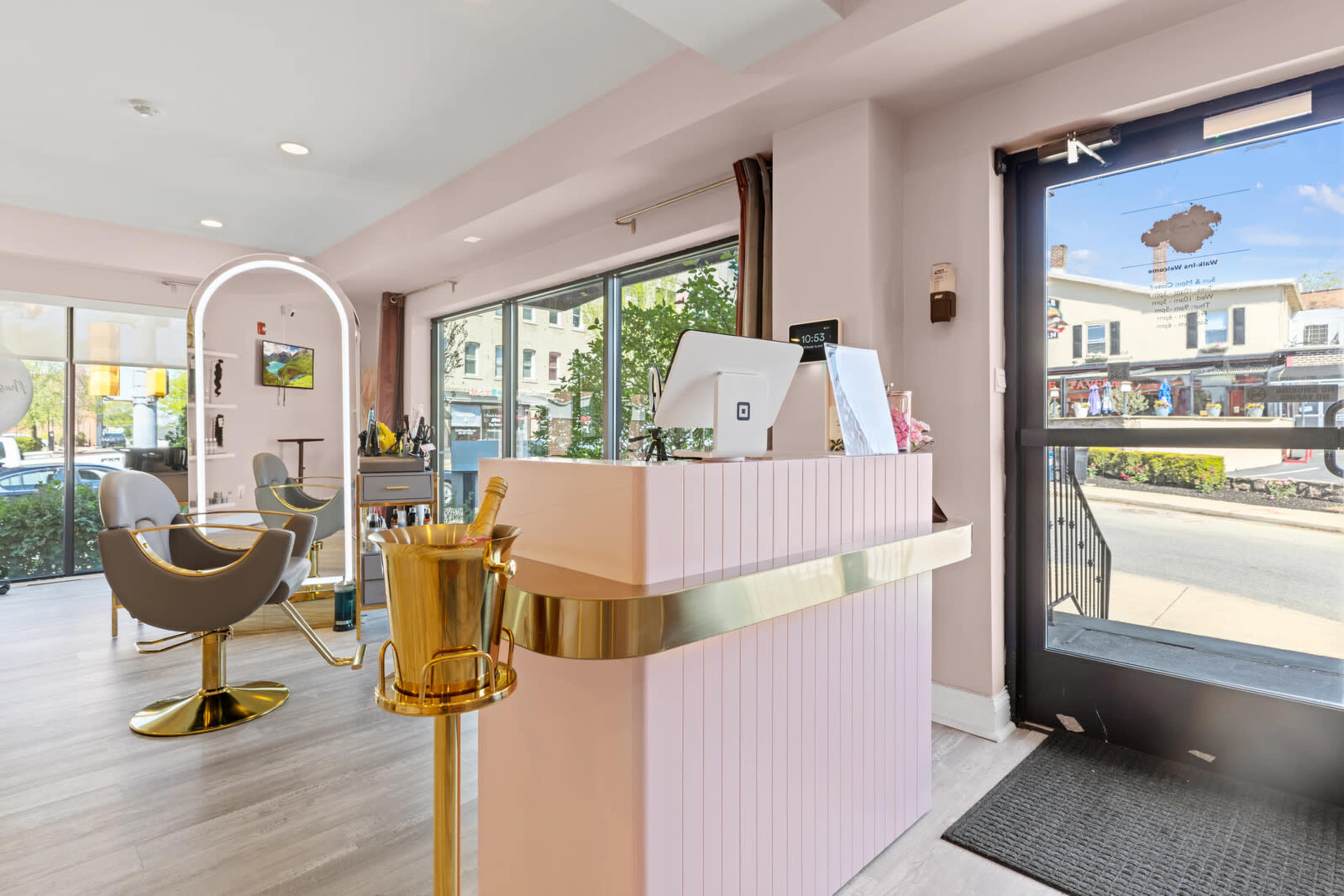 A modern beauty salon interior with a reception desk and styling chairs, featuring large windows that open to a street view.