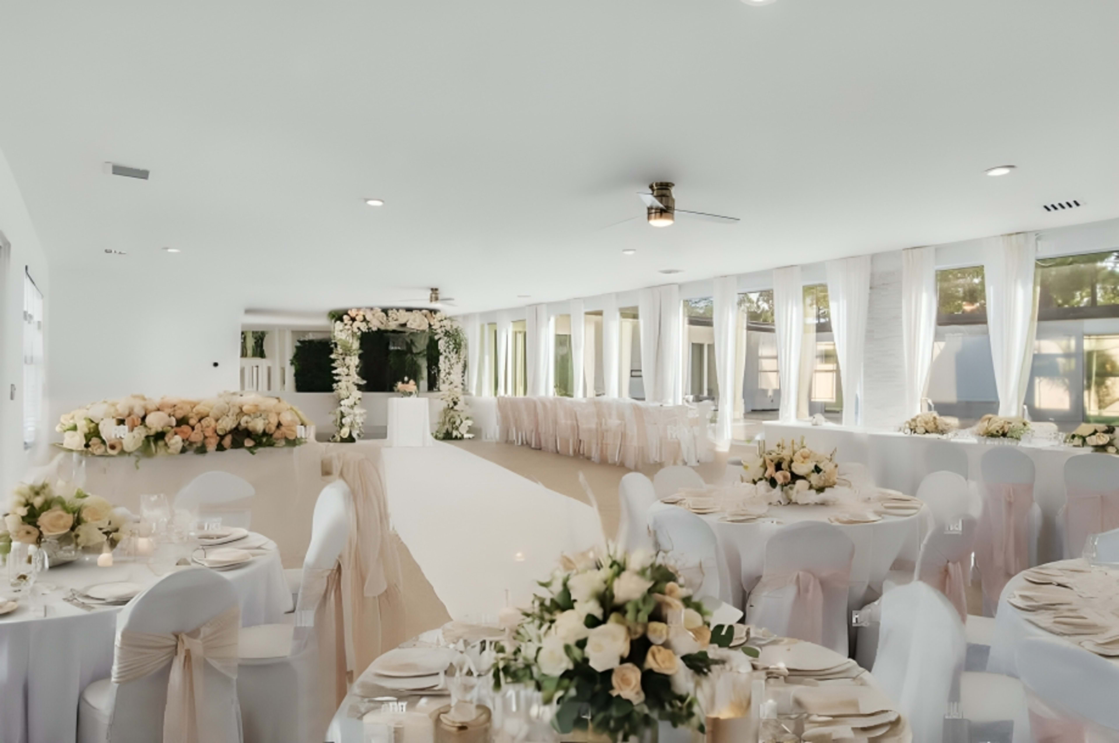 The image shows a spacious event venue set up for a celebration, featuring elegant round tables adorned with floral centerpieces and a decorated presentation area at one end.