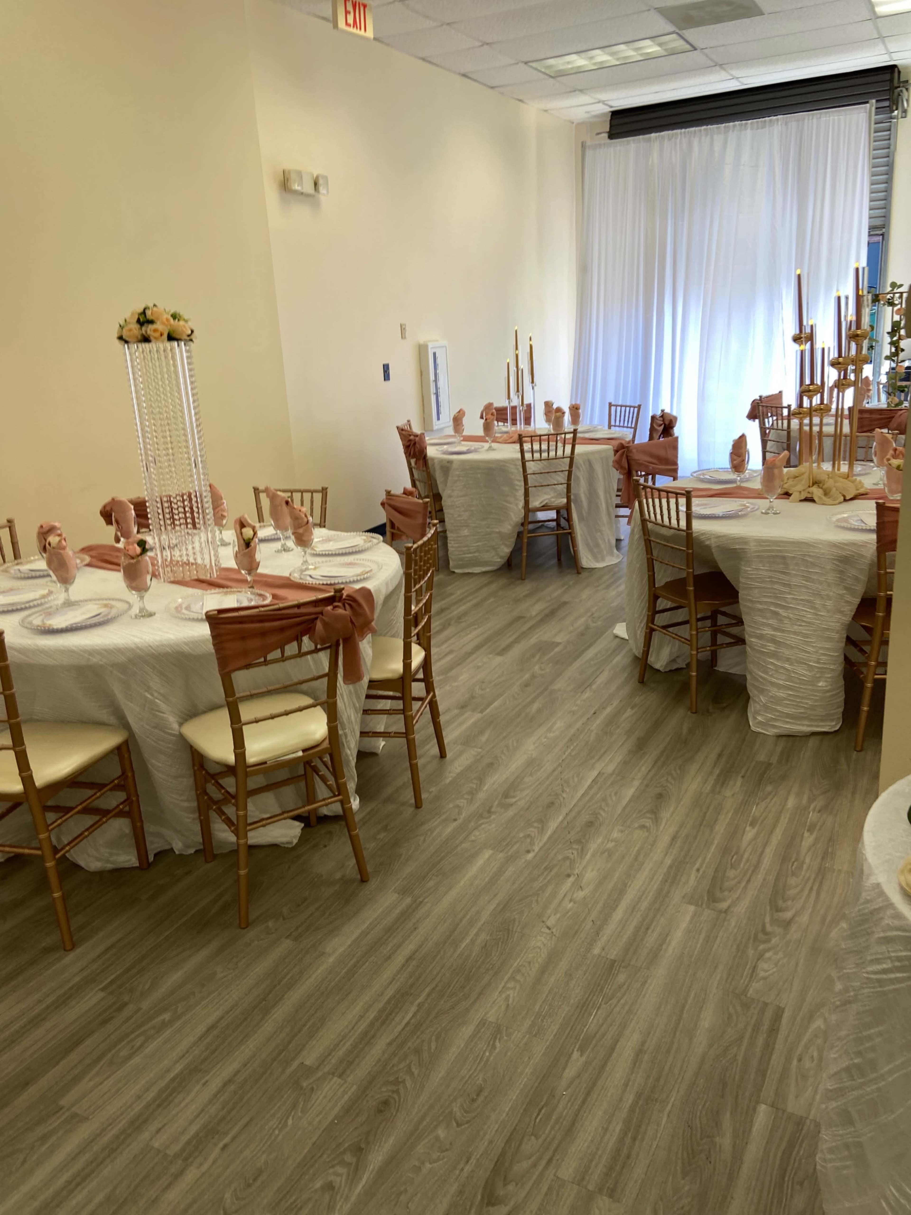 The image shows a banquet hall arranged with round tables covered in white tablecloths, adorned with decorative centerpieces and chairs set with light brown cushions.