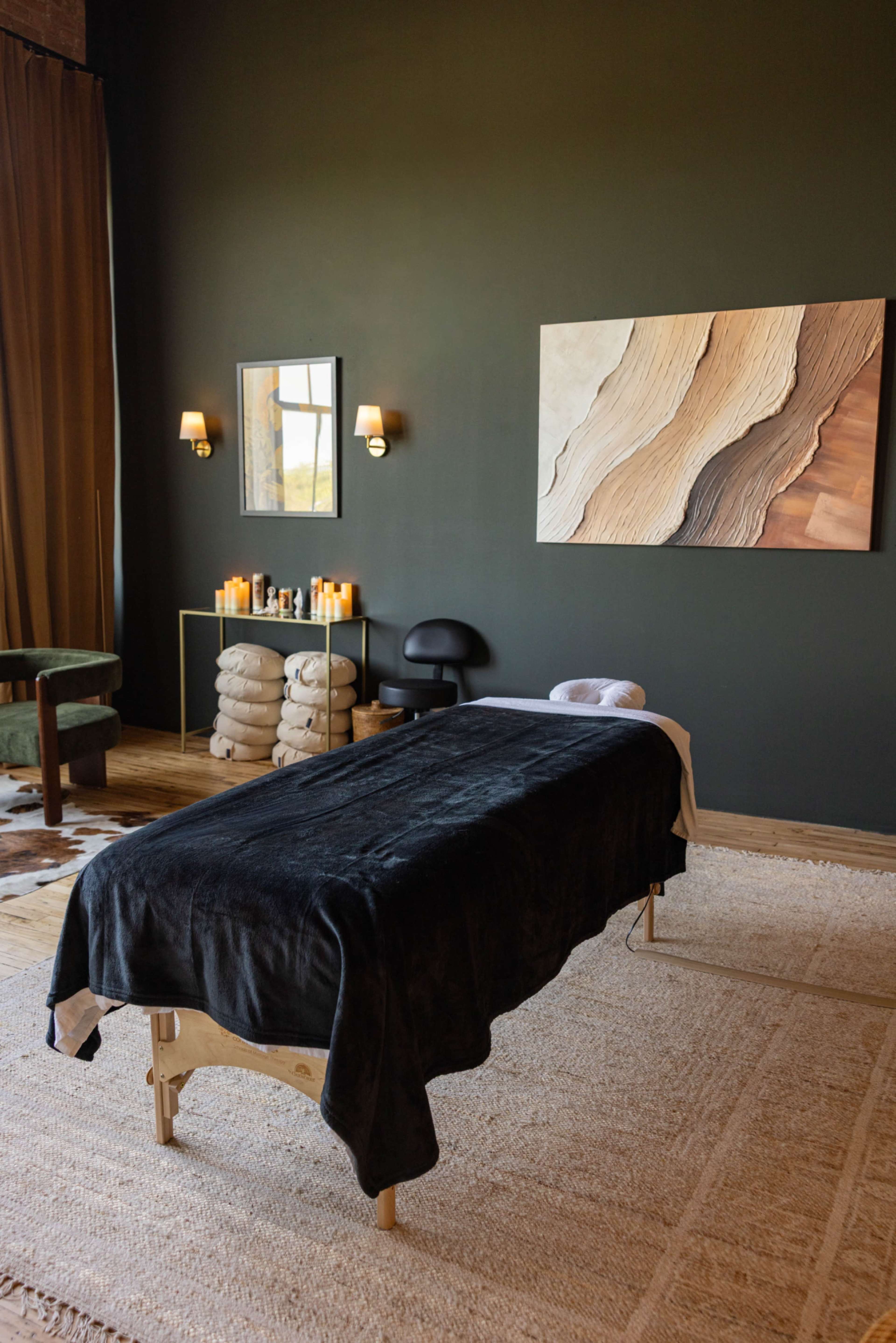 The image shows a serene massage room with a treatment table, neatly arranged towels, ambient lighting, and a piece of abstract wood-themed artwork on the wall.