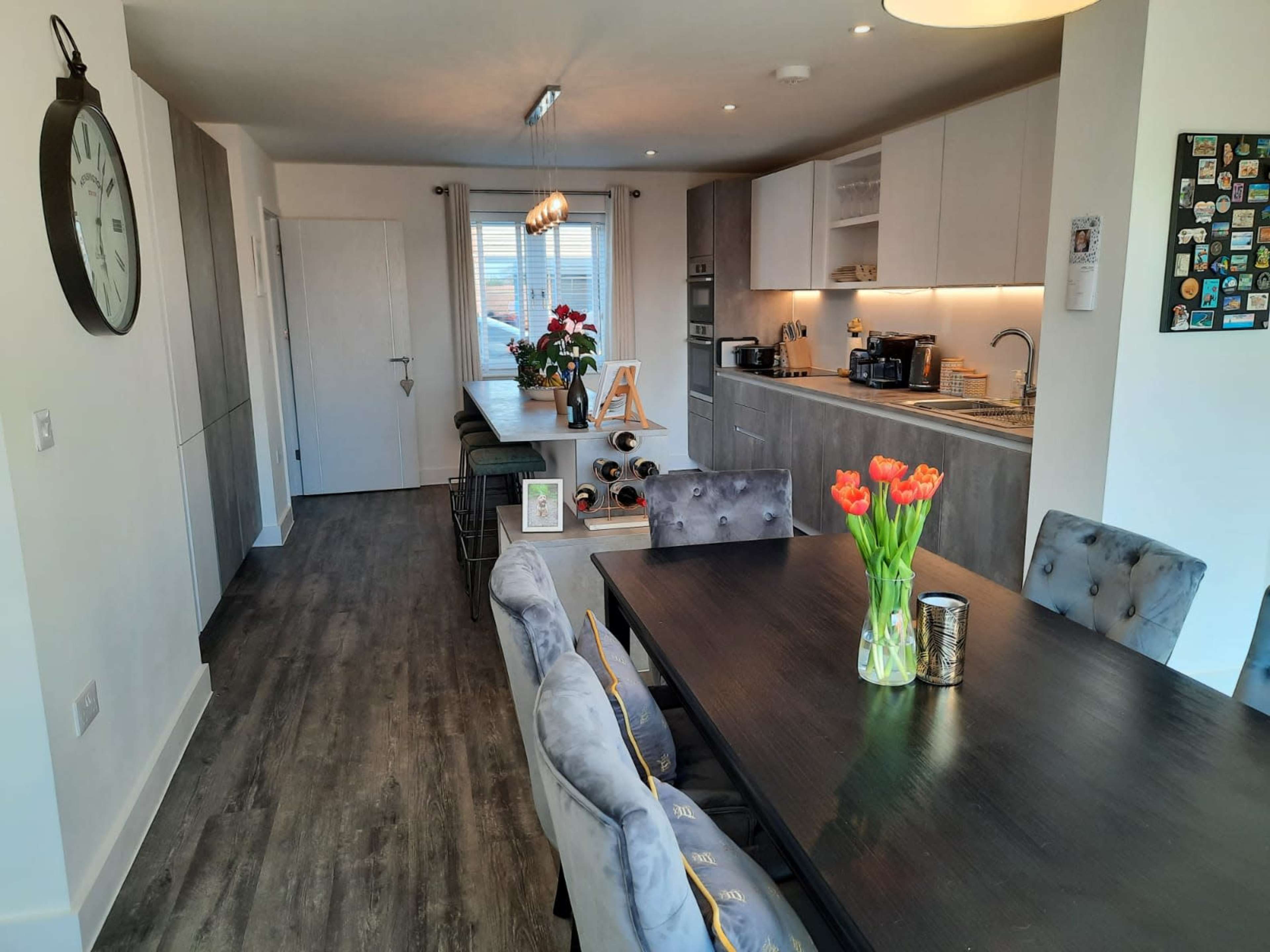 A modern kitchen and dining area feature a long table with flowers, gray cabinetry, and a wine rack.