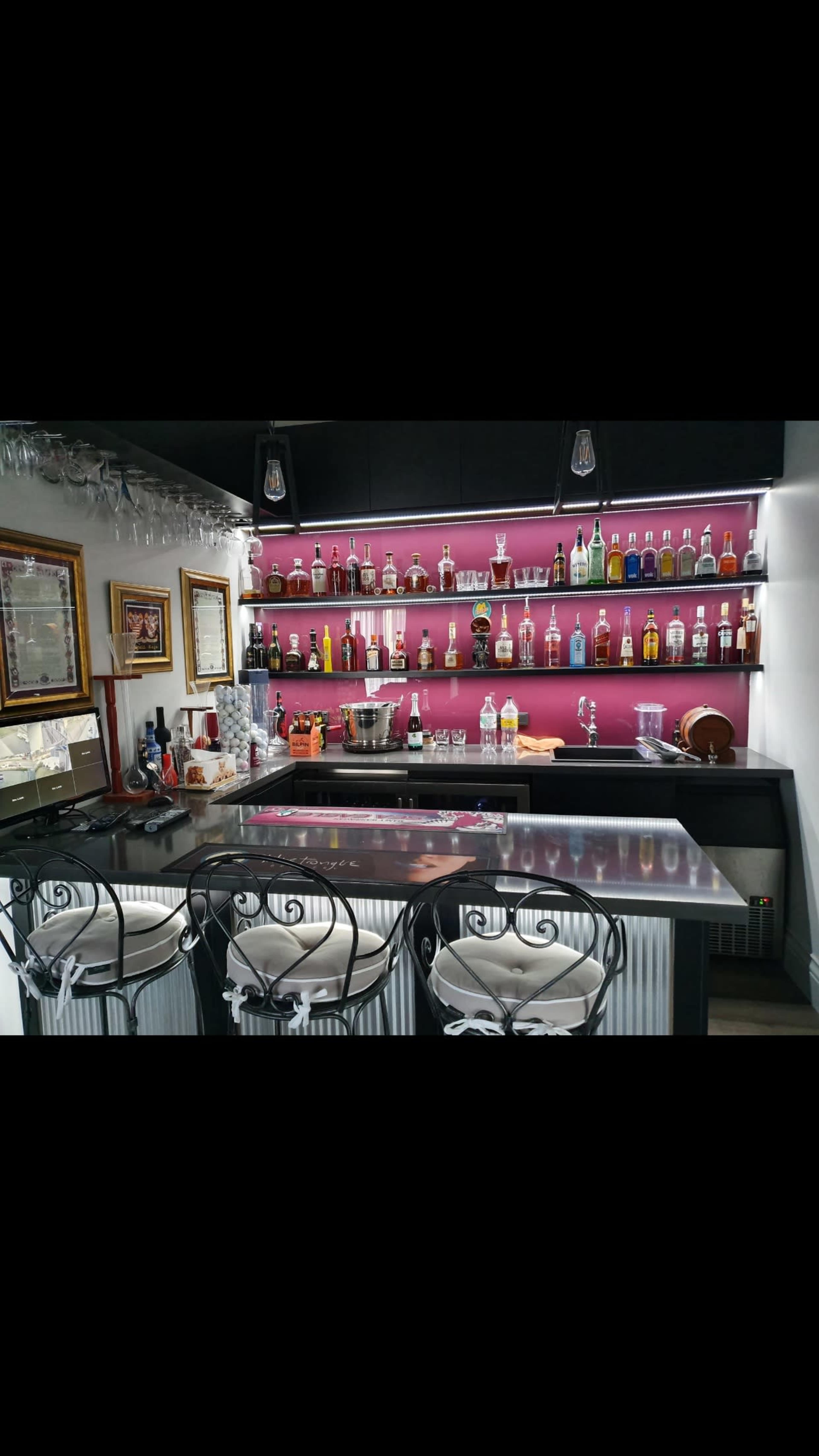 A well-stocked home bar featuring a glossy countertop, three metal-framed stools, and an illuminated shelf lined with various bottles.