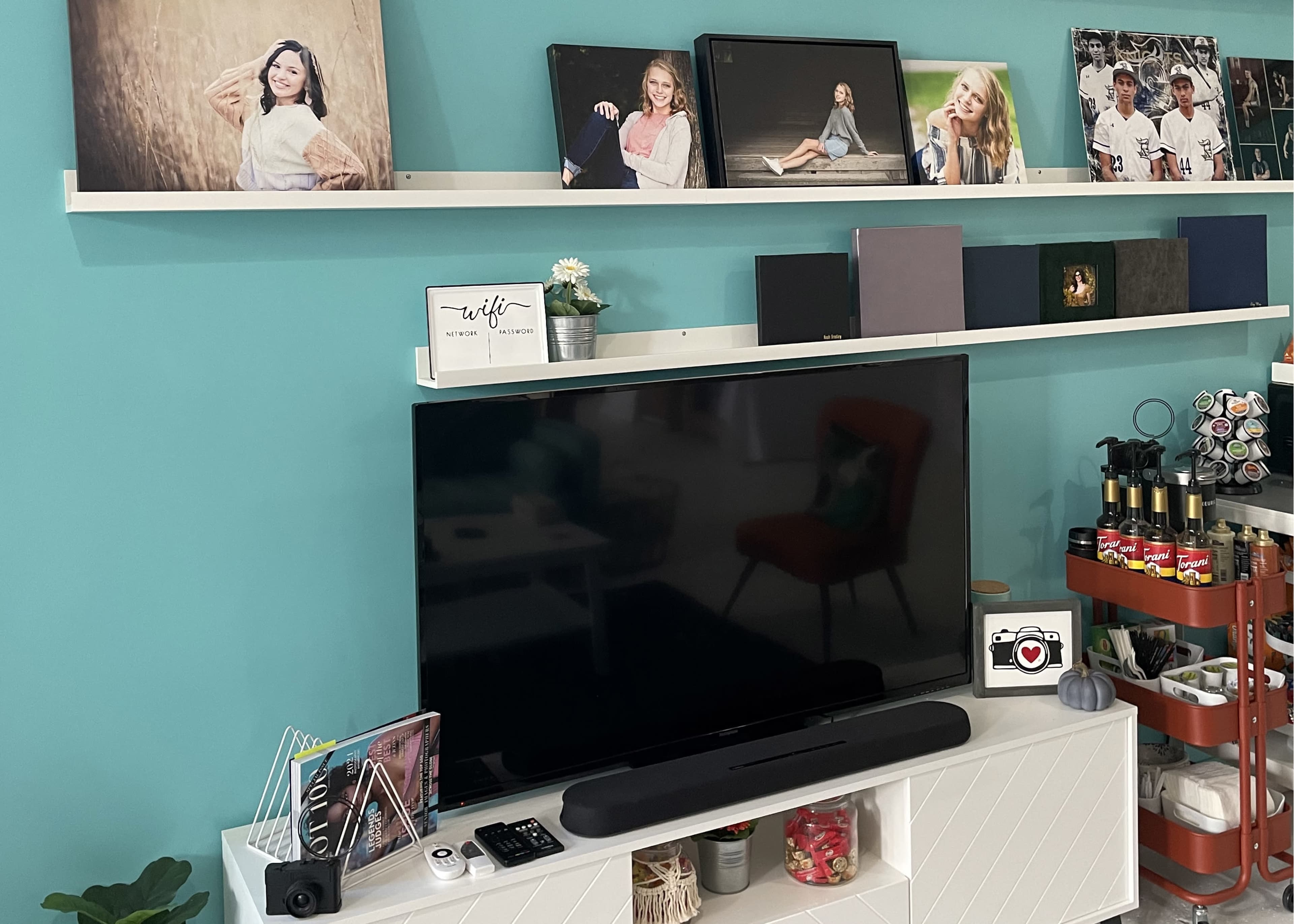 The image shows a living room area featuring a mounted TV on a white cabinet, with a shelf above displaying framed photographs against a turquoise wall.
