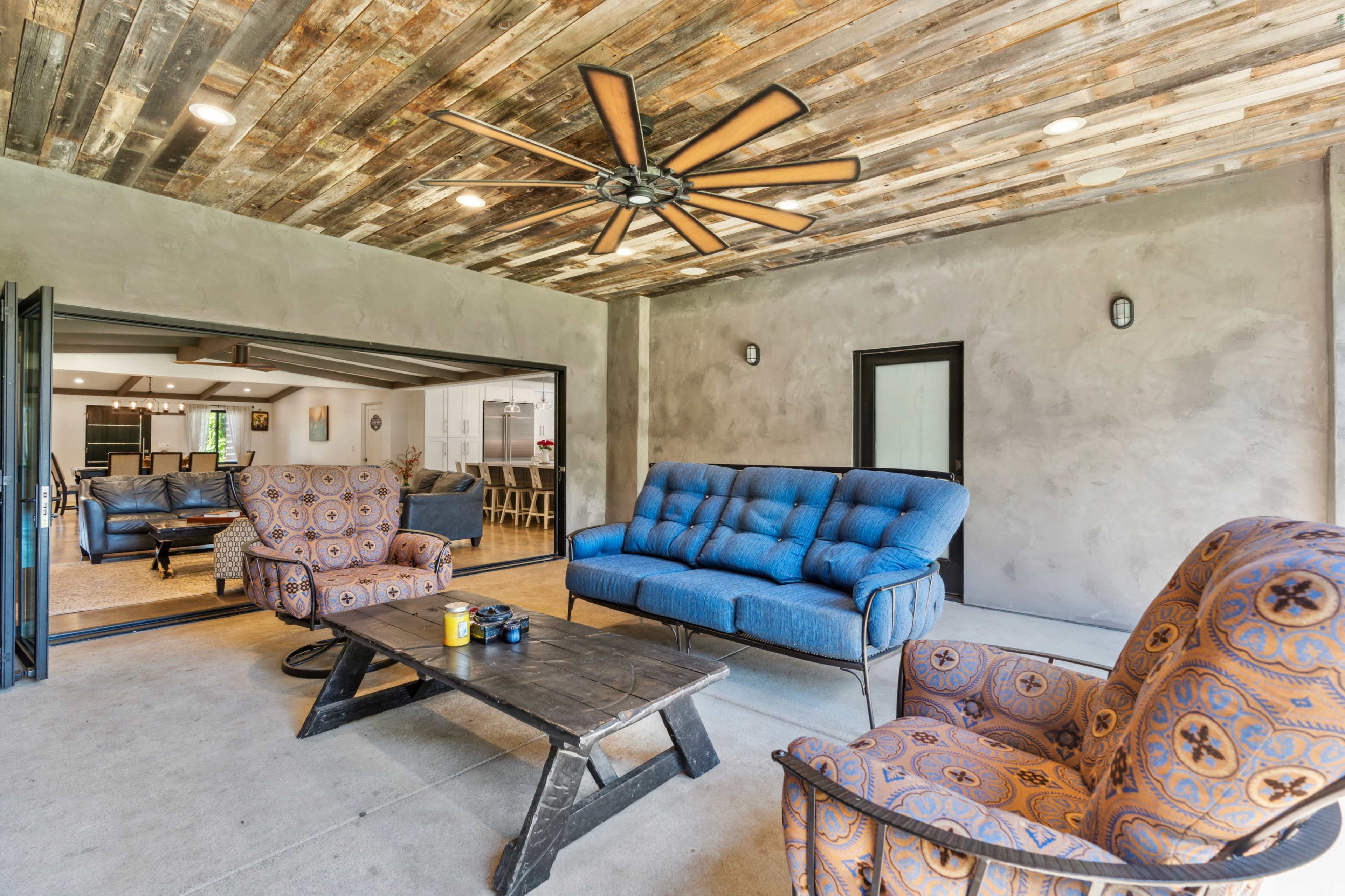 A cozy outdoor living space with a blue sofa and two patterned chairs arranged around a black wooden coffee table under a ceiling fan.