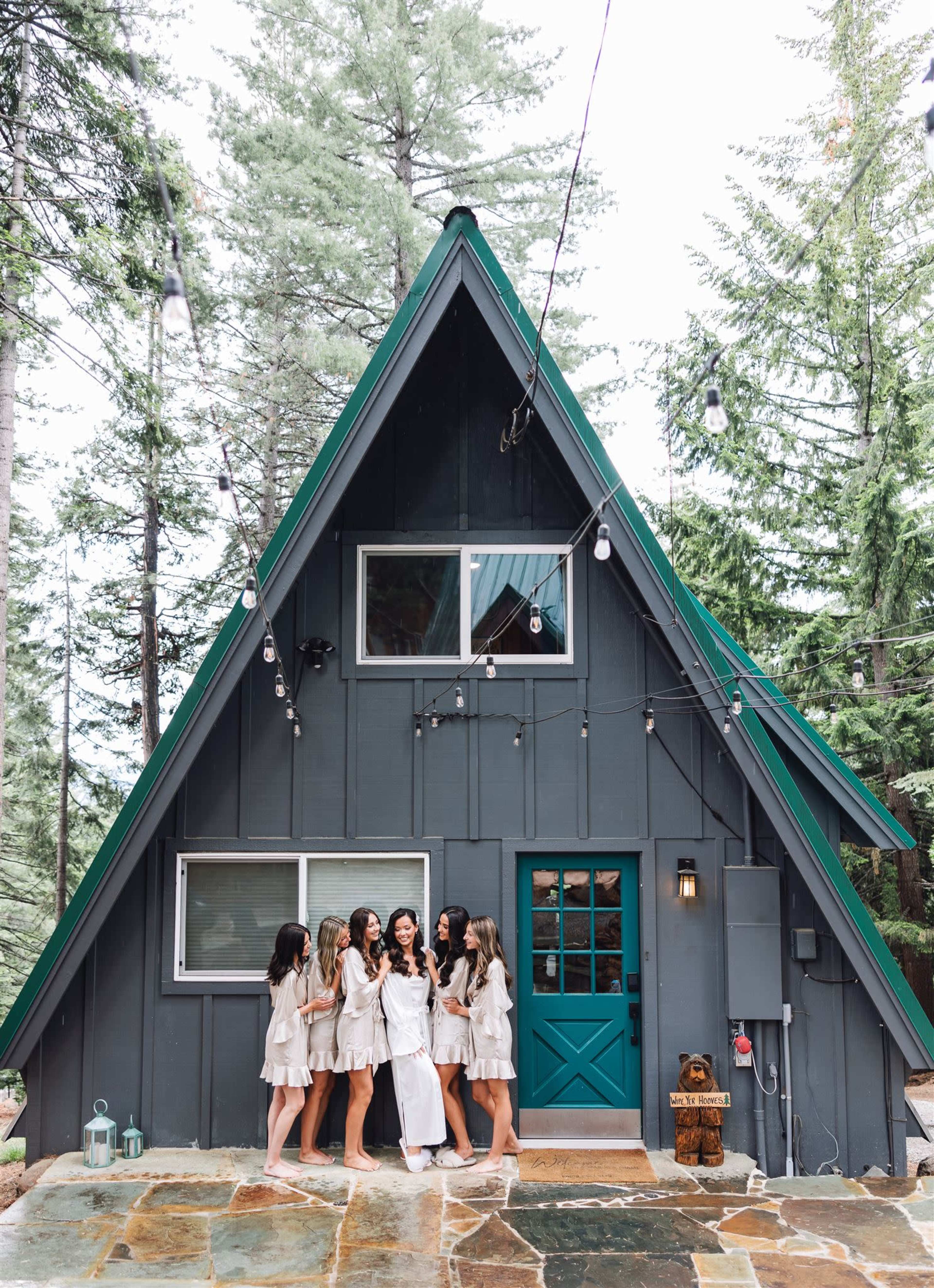 A group of six women in white dresses stands in front of a dark A-frame cabin surrounded by tall trees.