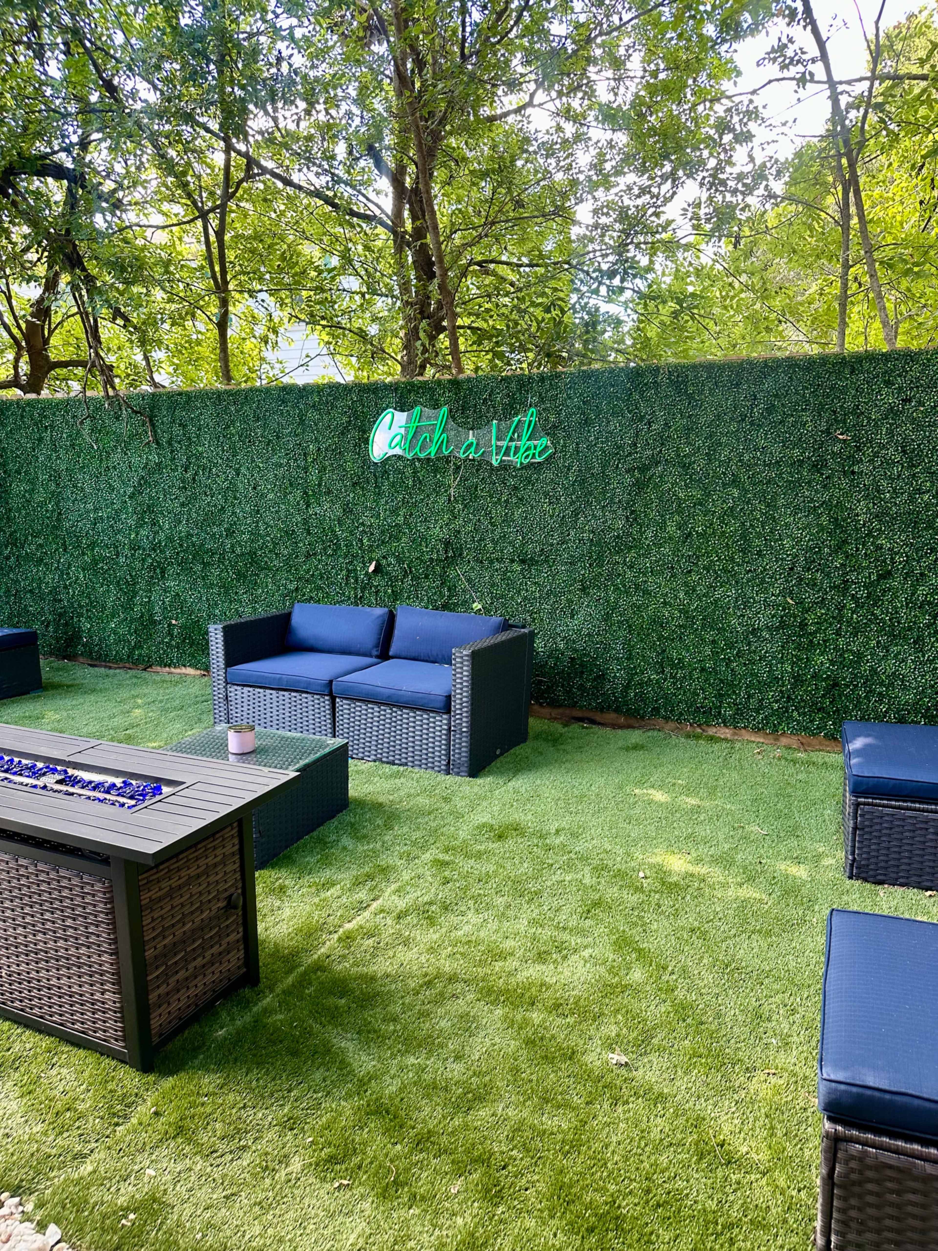 A seating area with dark wicker furniture is set against a green hedge featuring a neon sign that says "Catch a Vibe."