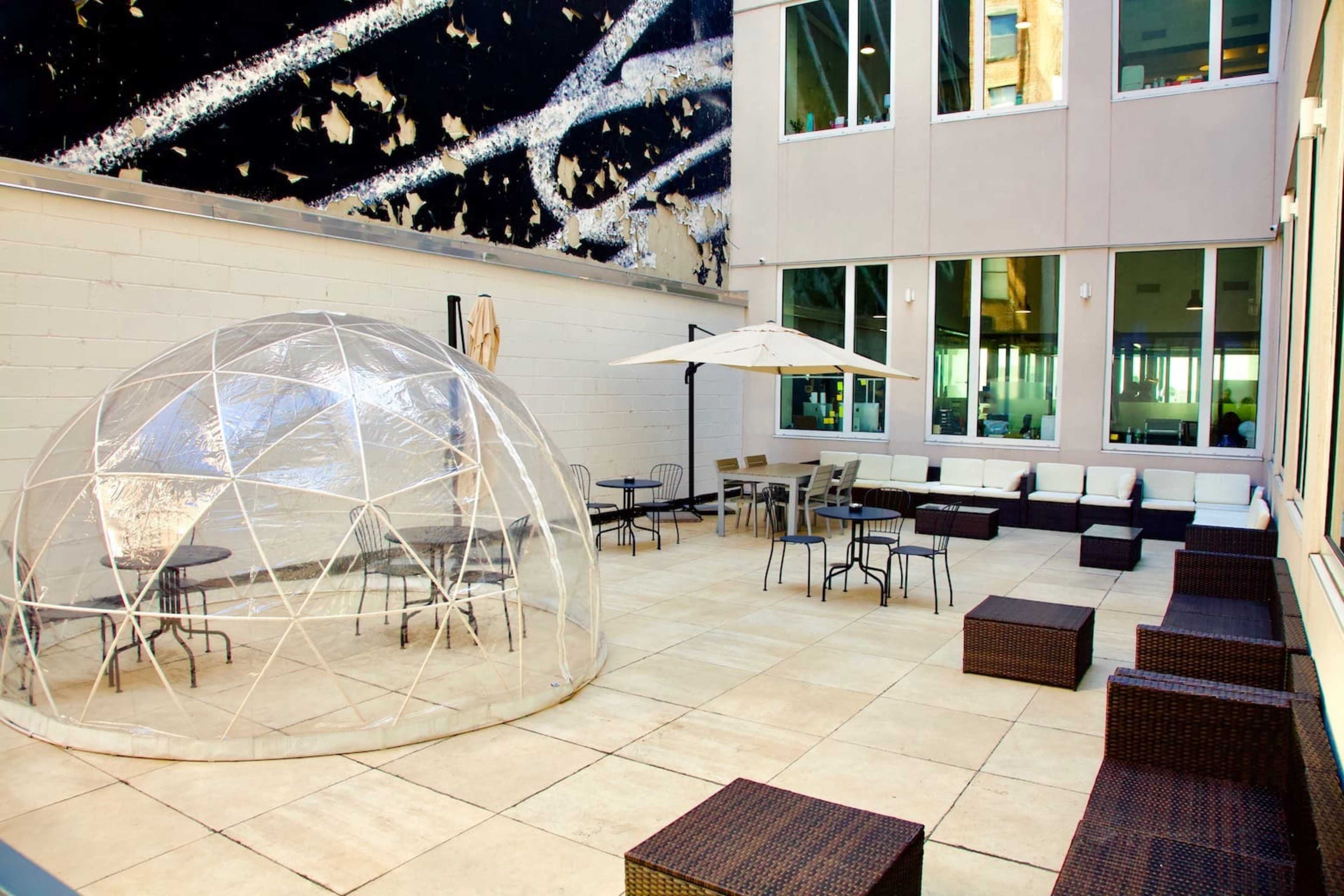 The image shows a modern outdoor patio area featuring a clear geodesic dome, several seating arrangements with dark wicker furniture, and a large umbrella.