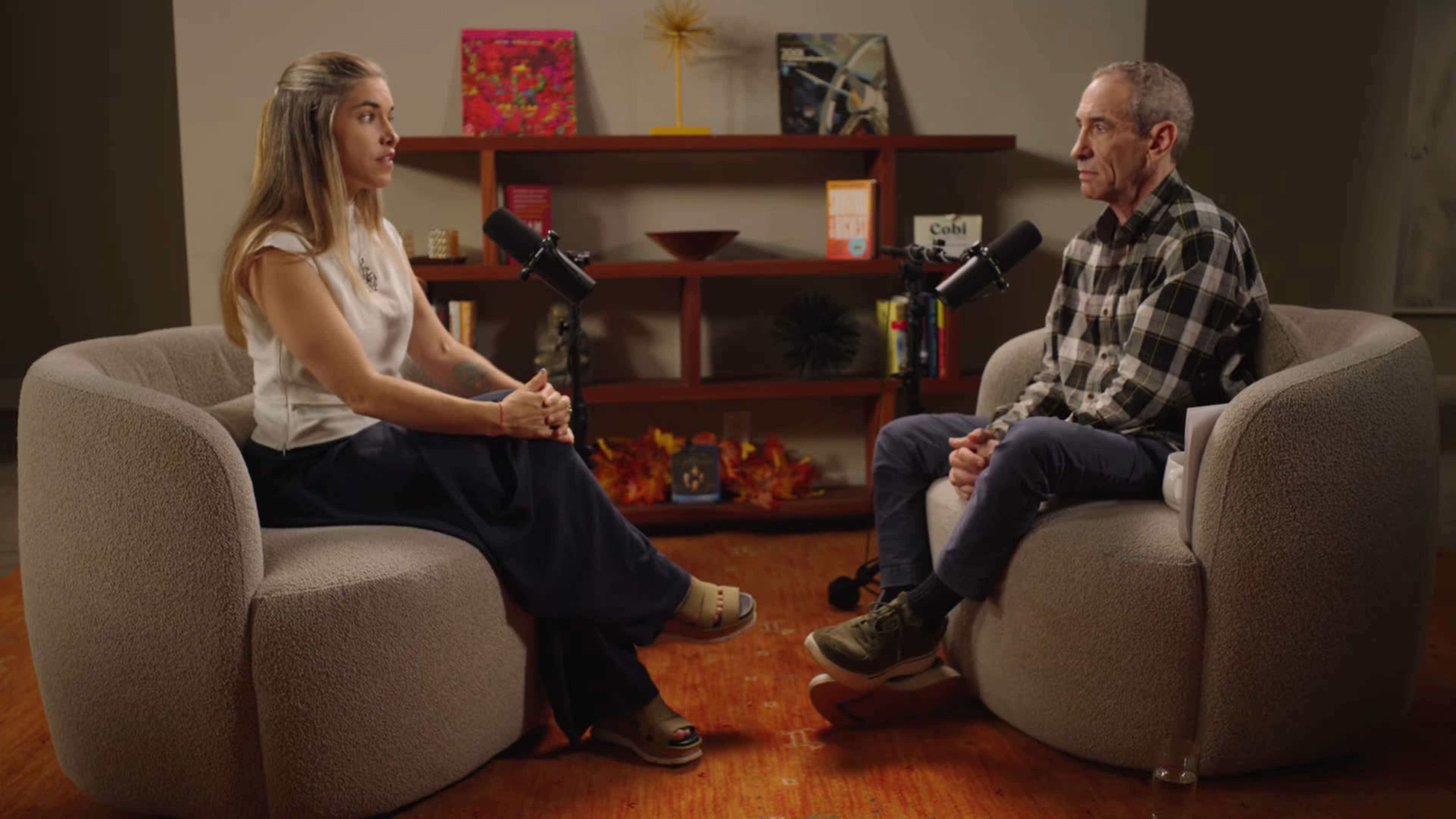 Two individuals are seated in a conversational setup on oversized chairs, each facing the other, with microphones in front of them and a shelf decorated with various items in the background.