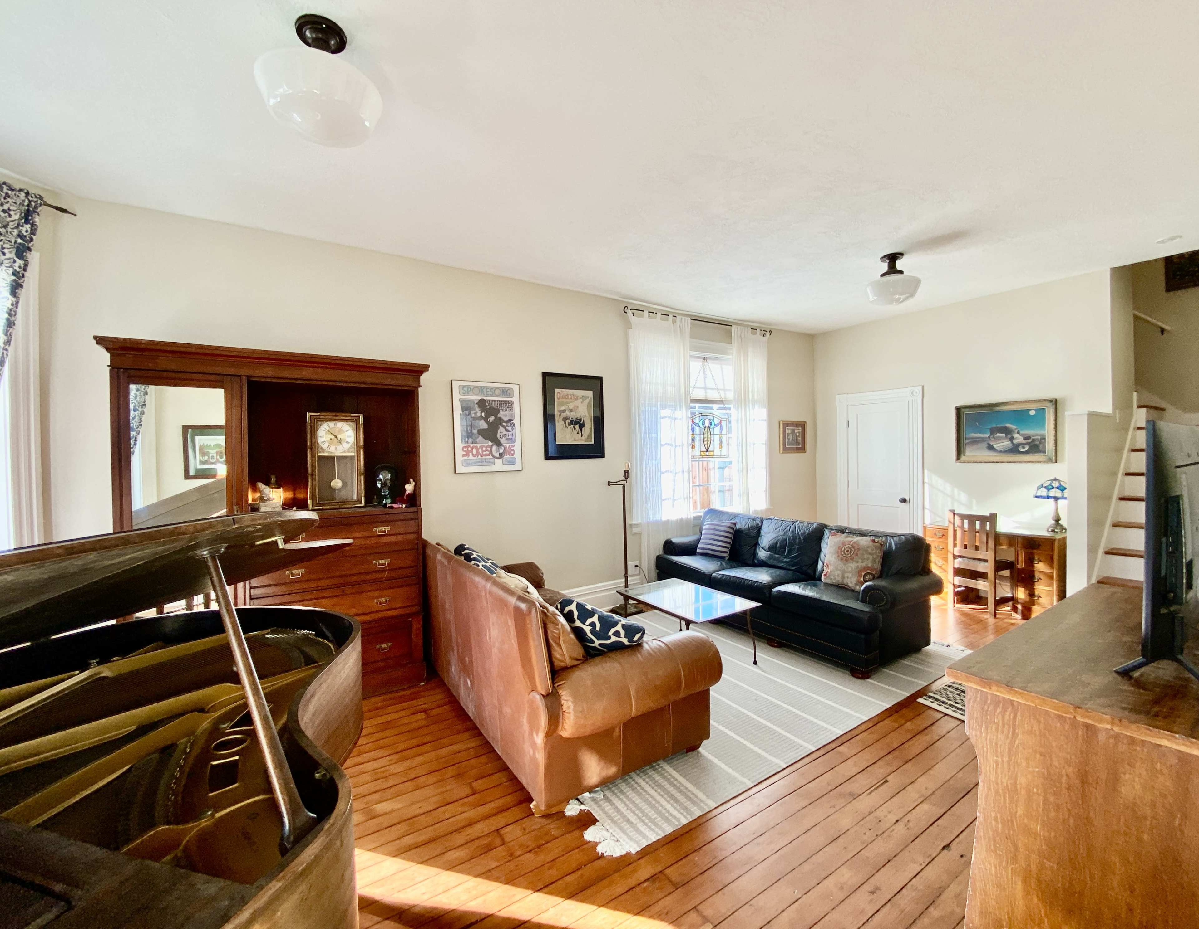 The image shows a bright living room featuring a piano, two sofas, a coffee table, and a wooden cabinet against a wall with several windows.