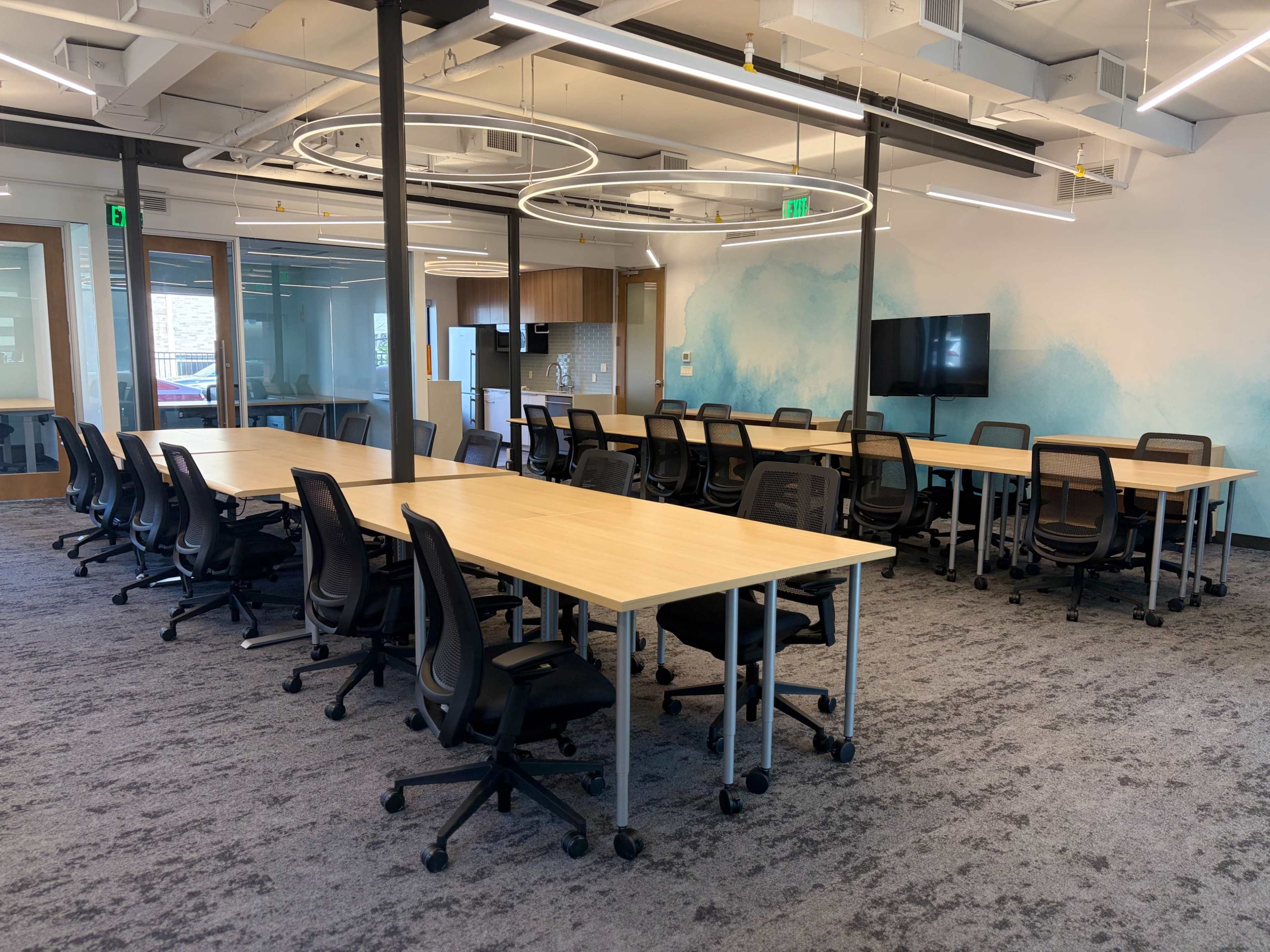 The image shows a modern office space with multiple tables and chairs arranged in two sections, along with a large screen on the wall and green accents.