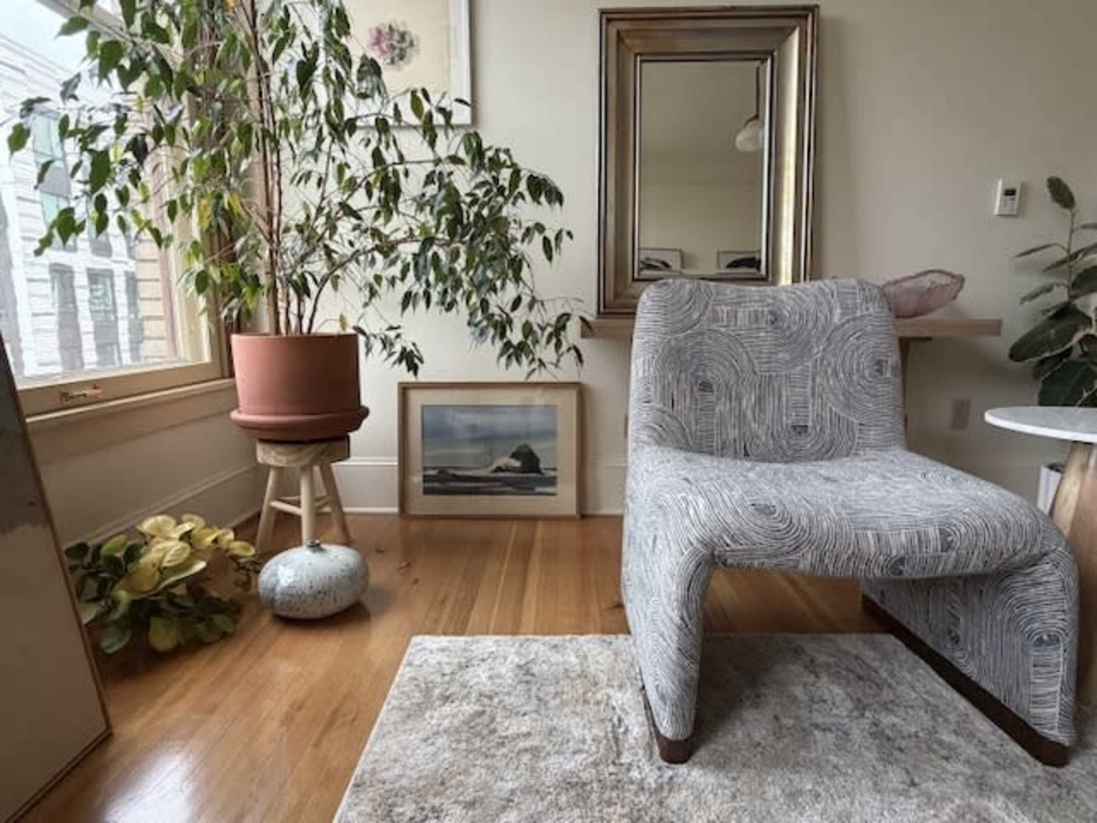 The image features a patterned chair next to a potted plant, a framed picture on the floor, and a round side table, all arranged in a well-lit room.