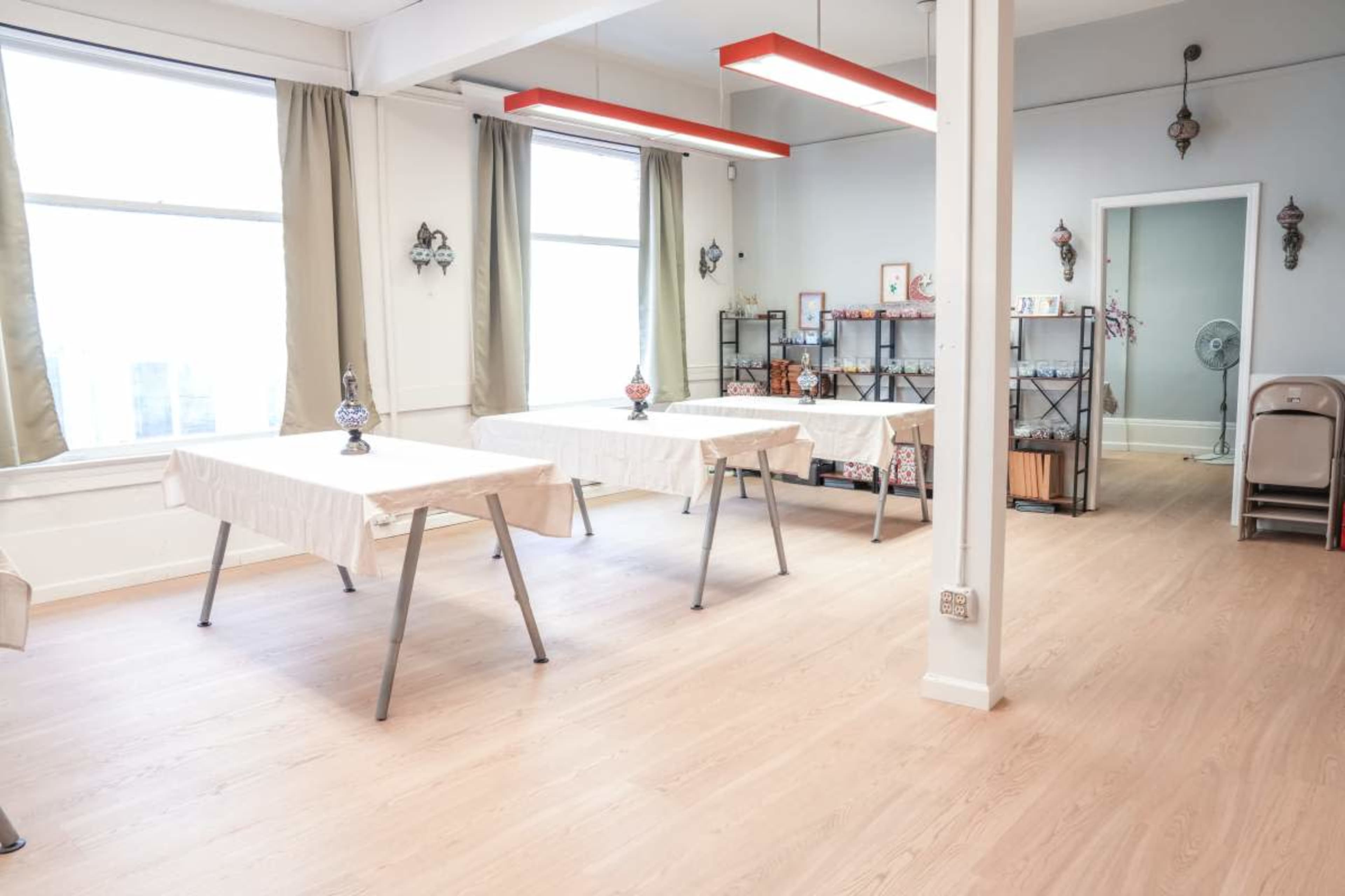 The image shows a light-colored room with two tables covered in white tablecloths and a few decorative items, along with a fan and shelving units against the walls.
