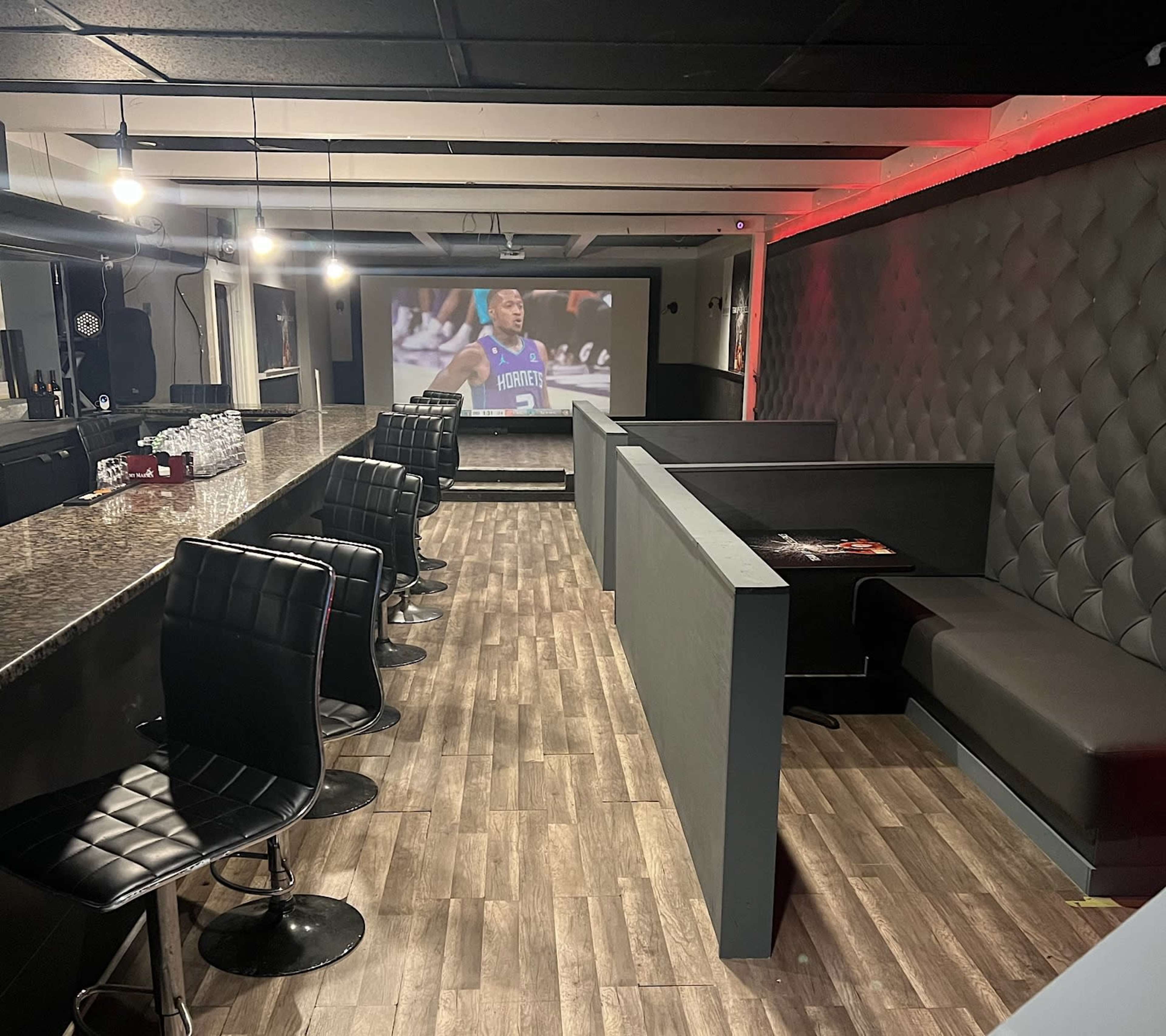 A modern lounge area with a bar, black leather chairs, and a screen displaying a sports event.