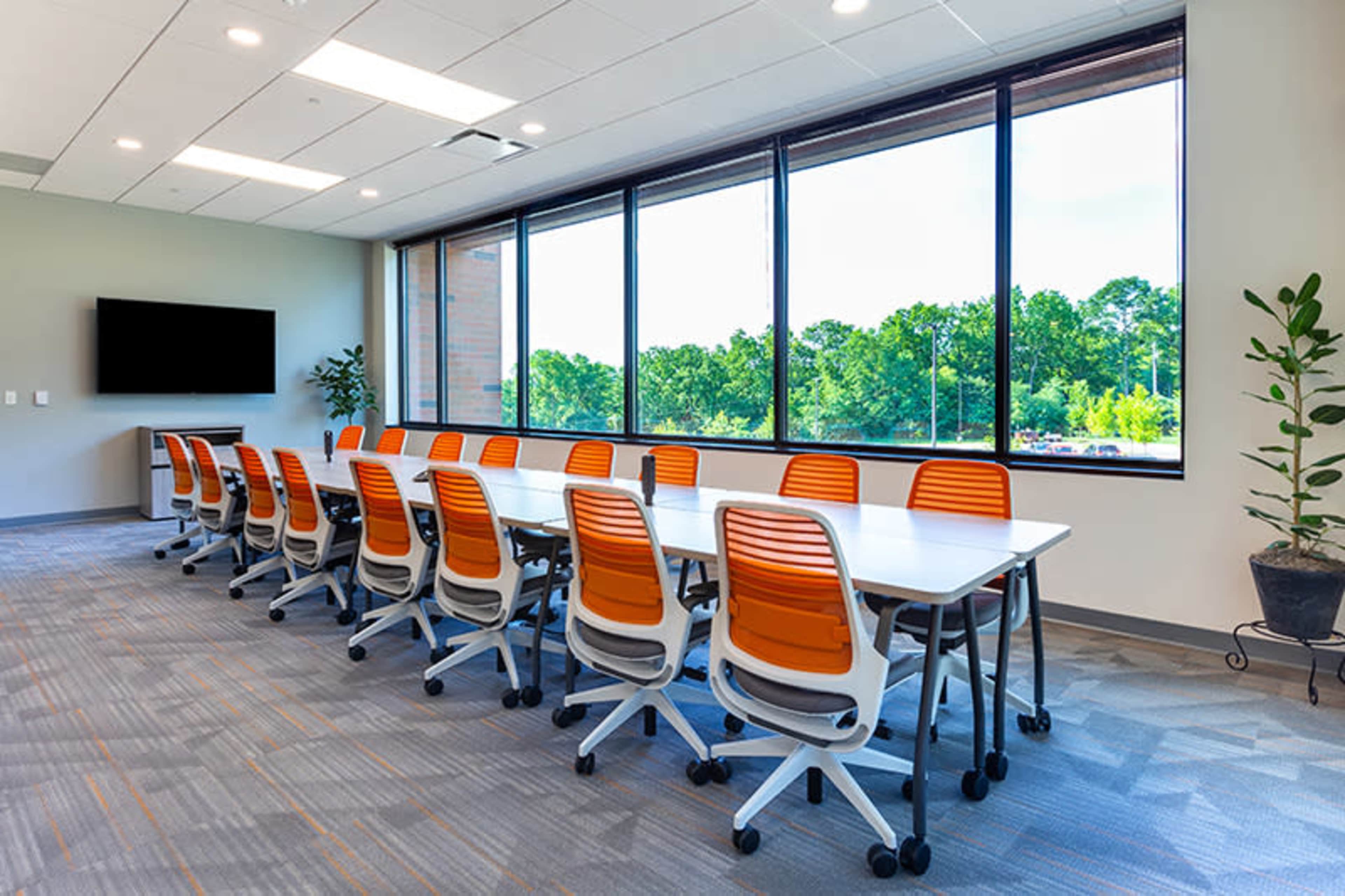 A large conference room with a long table and orange wheeled chairs is illuminated by natural light streaming through large windows overlooking green trees.