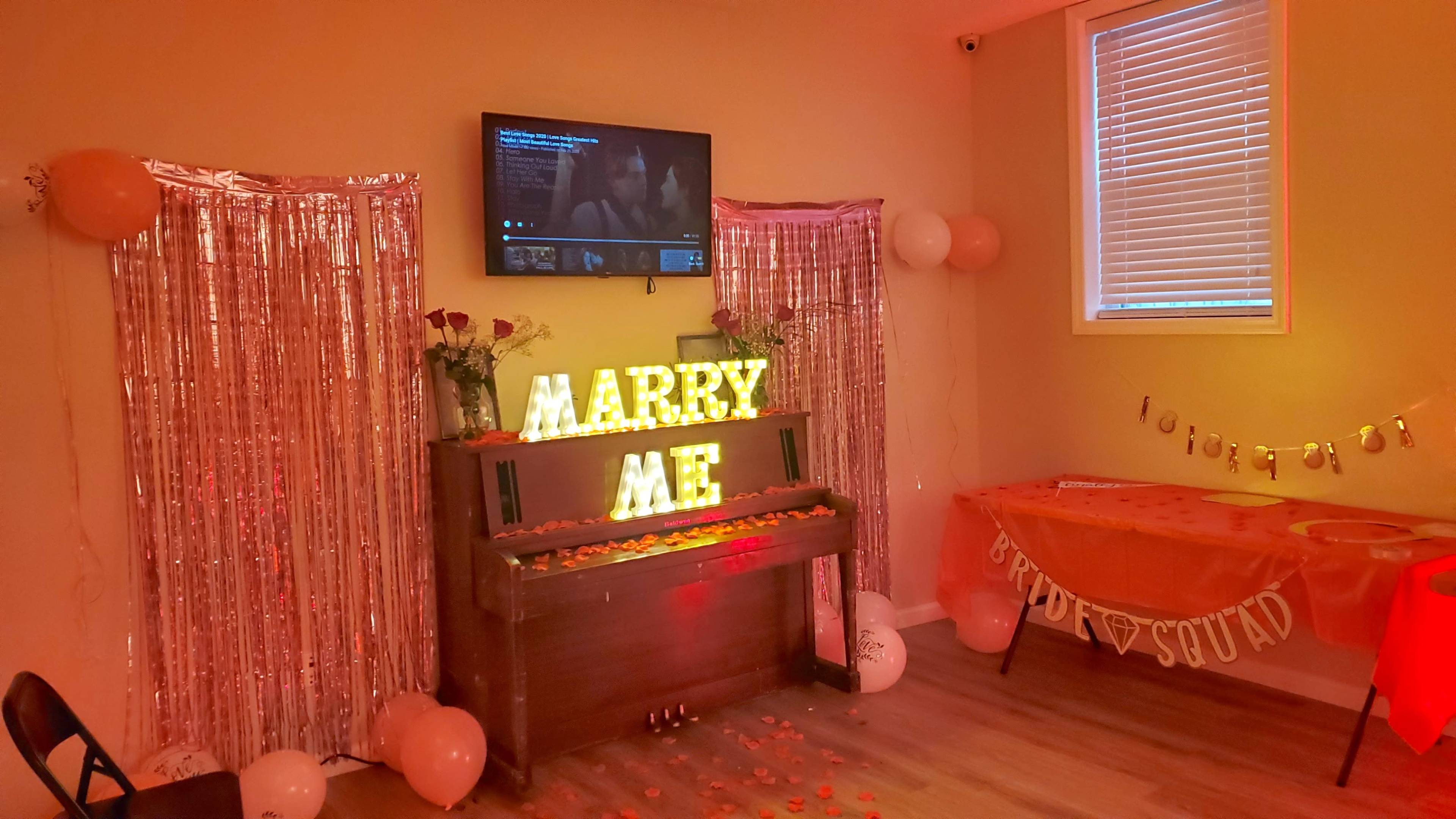 The scene features a small room decorated with orange lighting, a TV playing a video, a piano adorned with illuminated letters spelling "MARRY ME," and a table set with decorations for a celebration.