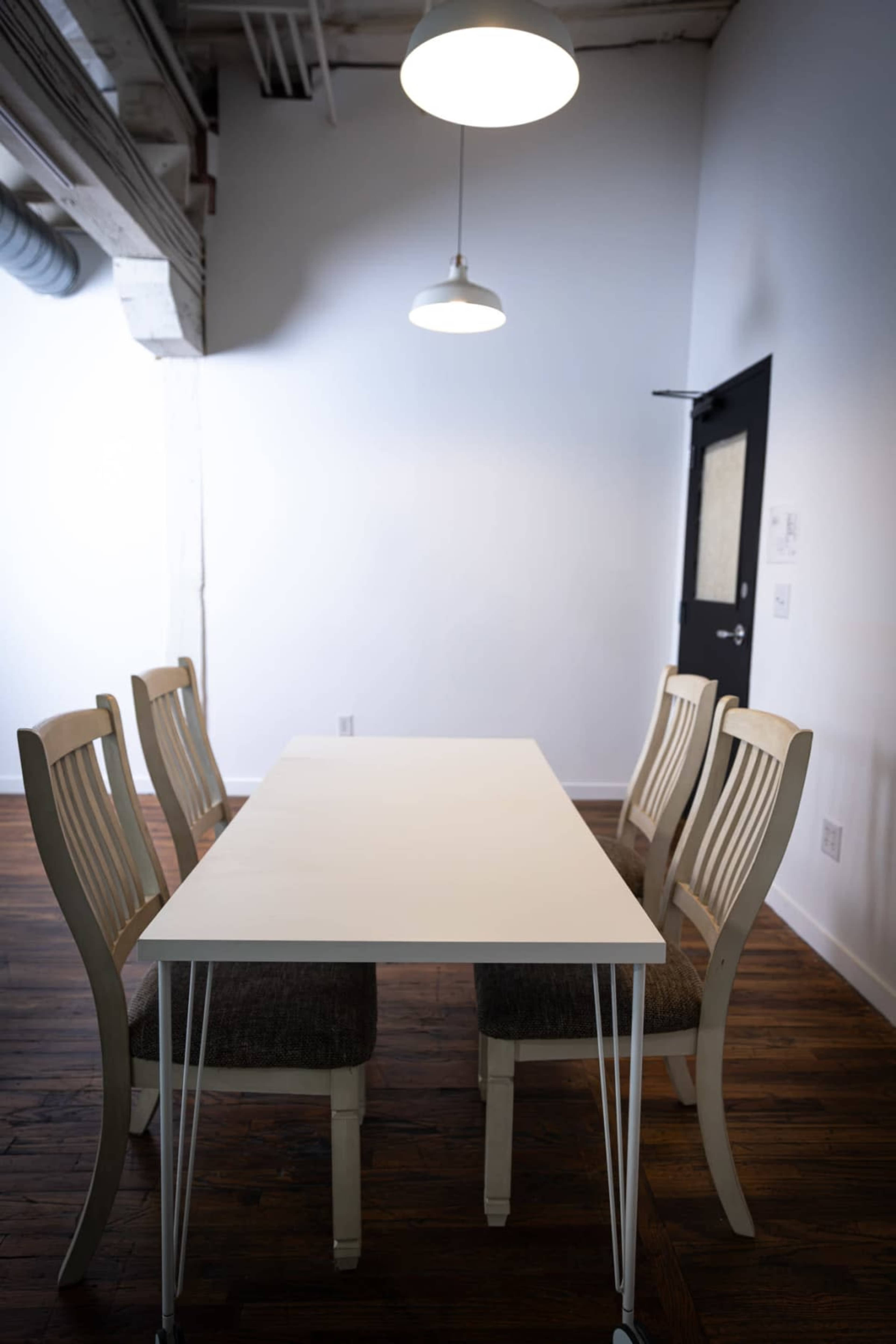 A long white table is set in a minimalist room with four wooden chairs arranged around it, illuminated by two pendant lights above.